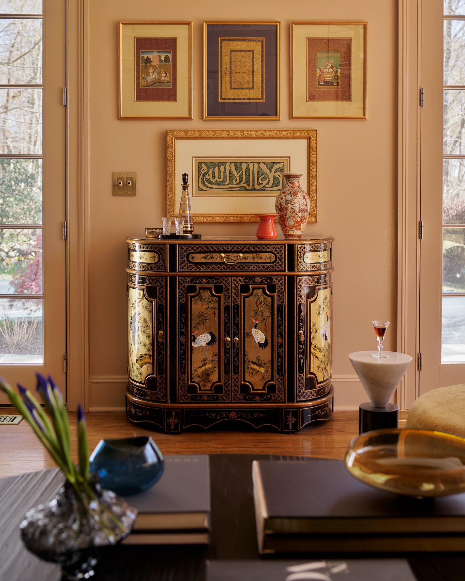 A lacquered chinoiserie console sets the stage for a salon vignette steeped in global influence. Framed Islamic calligraphy and Mughal miniatures form a curated salon-style gallery wall, anchoring the space with reverence and refinement. A mix of cer