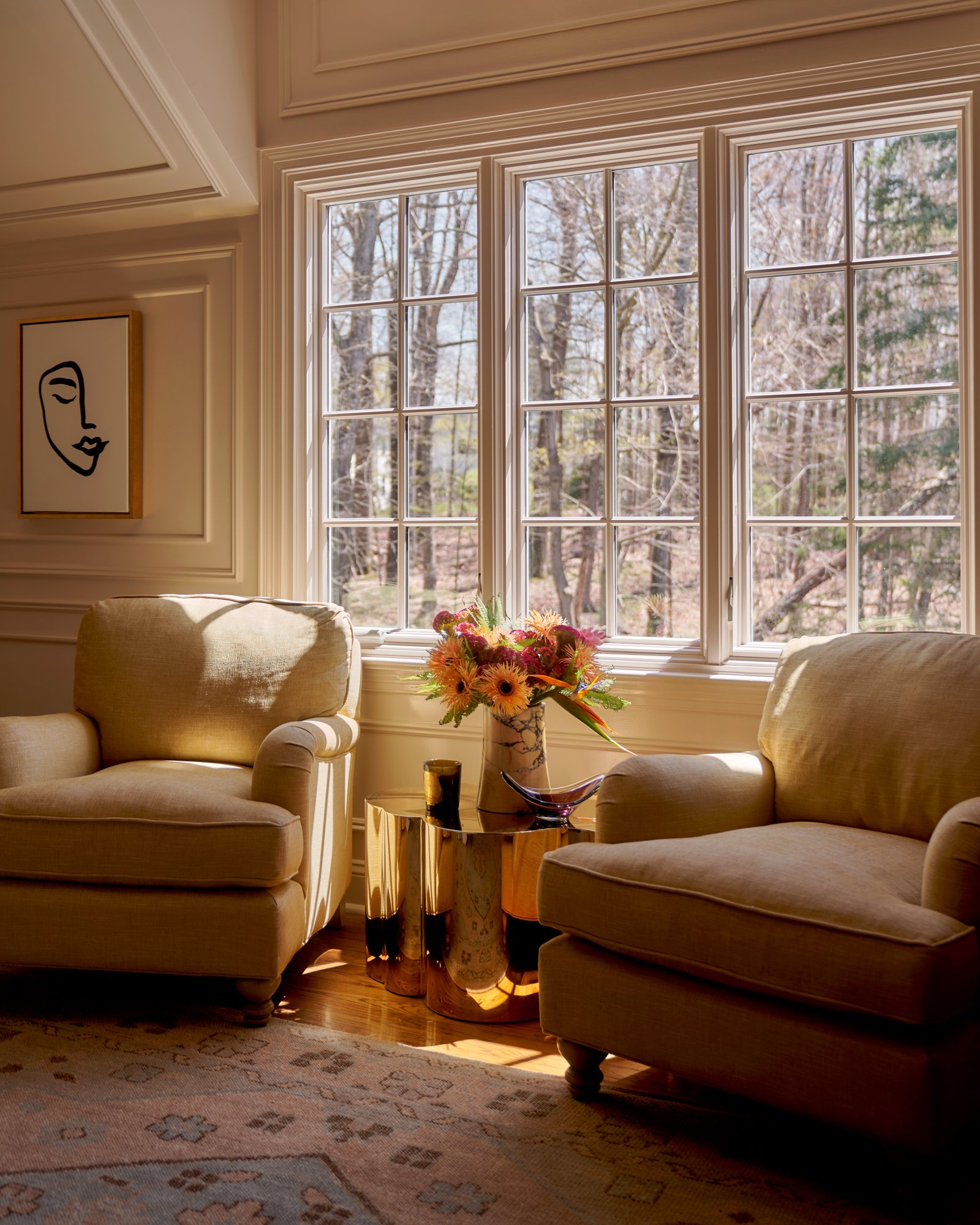 A sun-drenched moment of repose—this intimate sitting area balances modern softness with classic millwork and curated accents. Creamy linen club chairs flank a sculptural gold side table, while a single line drawing anchors the space with graphic ele
