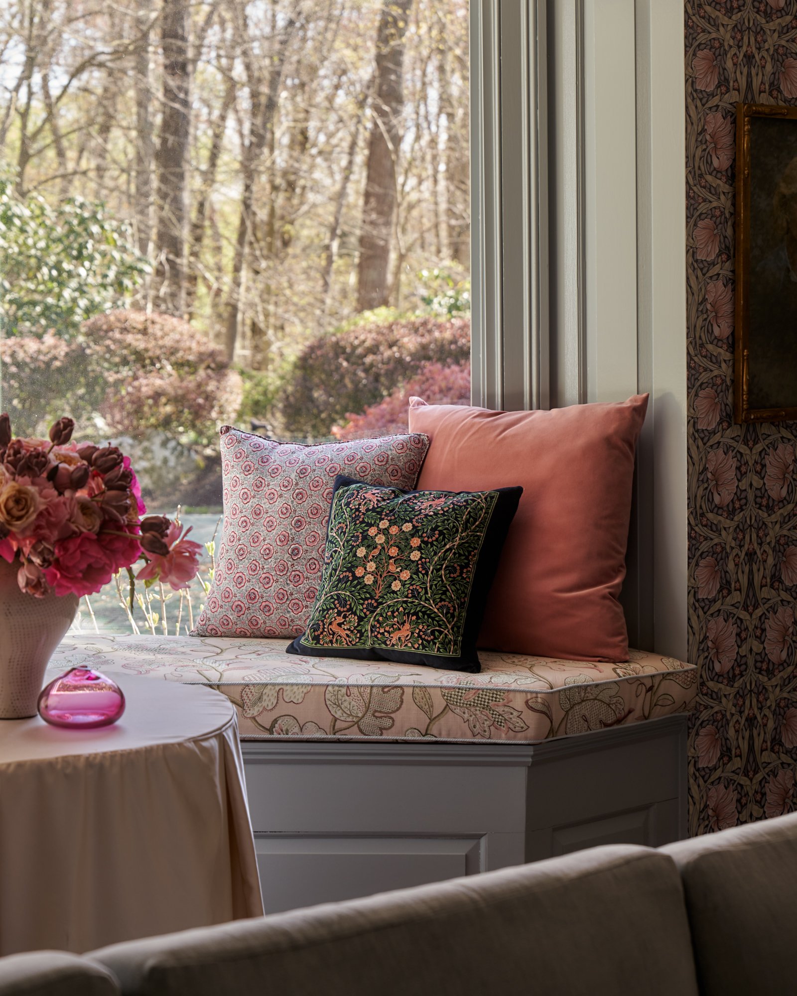 Nestled into the bay, this upholstered window seat becomes a romantic punctuation in the room’s floral narrative. Embroidered linen, velvet, and block-printed cushions mingle against a garden backdrop, blurring the line between inside and out. A gath