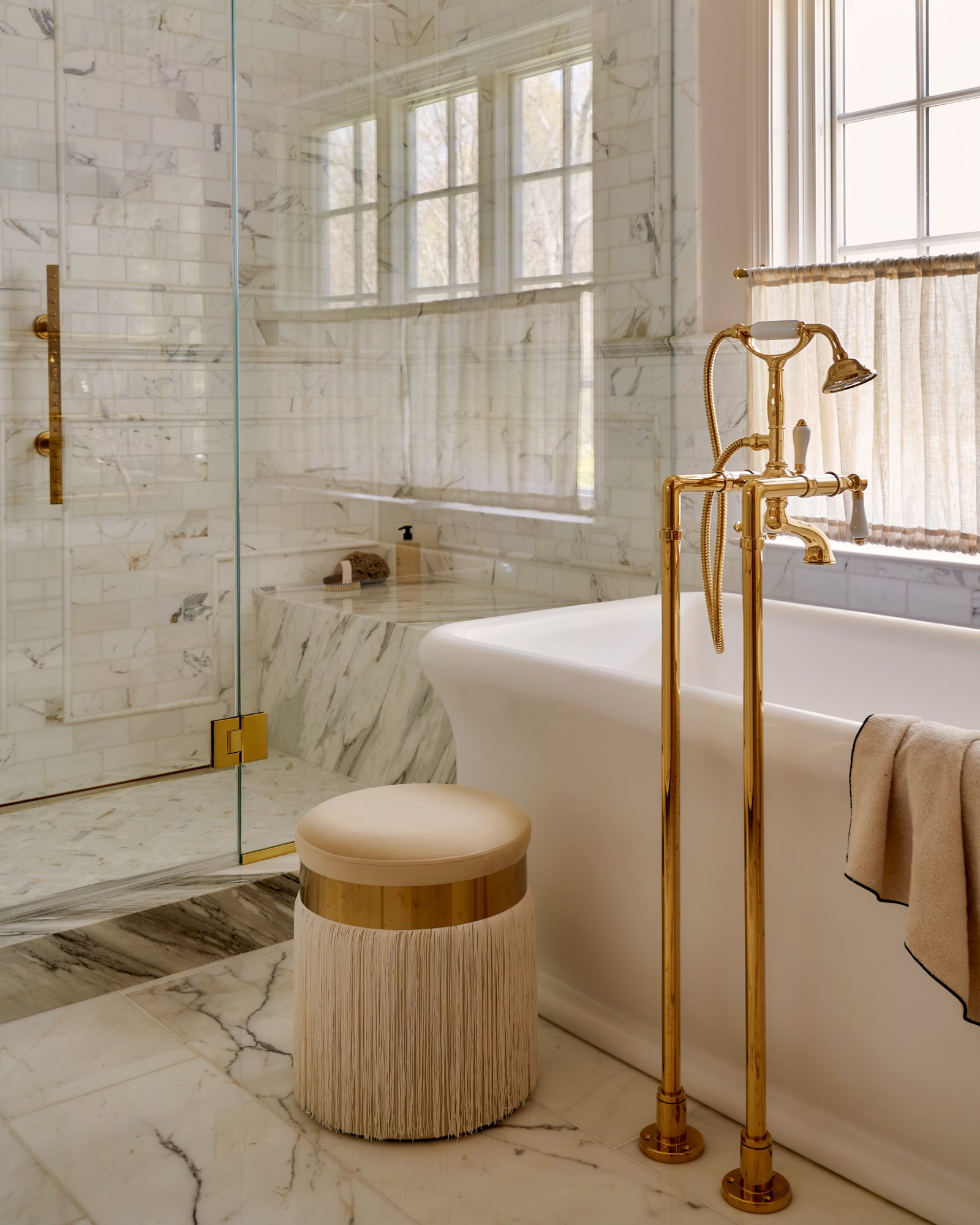 This primary bath is a masterclass in quiet luxury—wrapped in honed Calacatta marble with brushed brass fittings and a freestanding tub as sculptural focal point. A fringed vanity stool adds a whisper of vintage glamour, while sheer café curtains sof