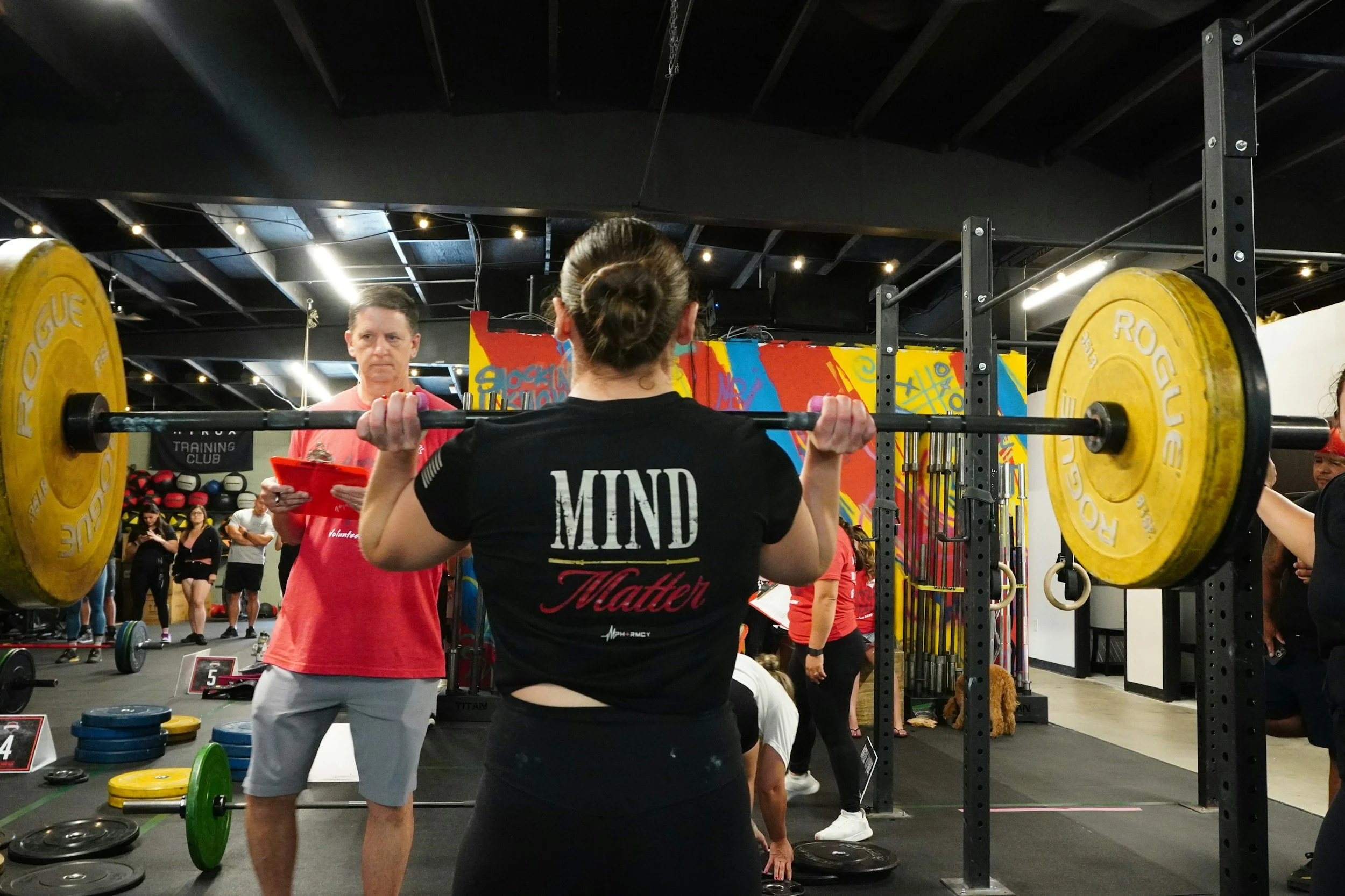 A woman shown from behind performs a barbell squat in a gym, with yellow weight plates on the bar and a behind-the-scenes view of a fitness class or event with multiple people, some lifting weights, and colorful workout equipment and mural in the background.