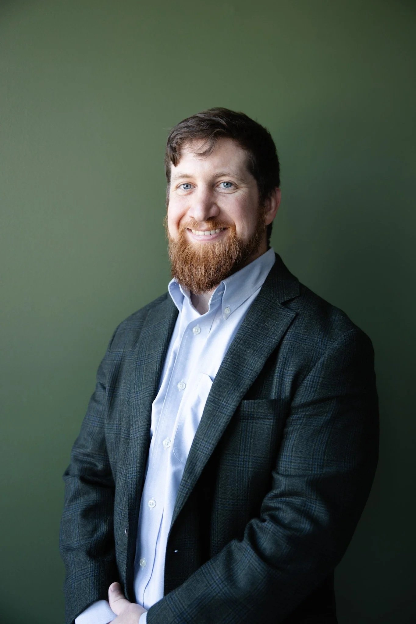 A man with light skin, brown hair, and a beard, wearing a dark checkered blazer and a white collared shirt, standing against a green background, smiling at the camera.
