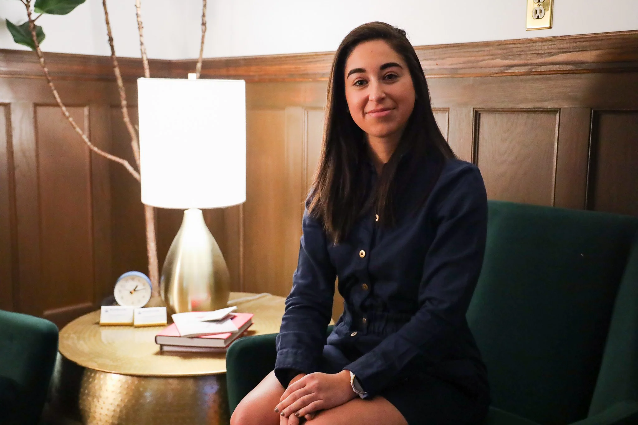 A young woman with dark hair and a slight smile, sitting in a room with wood-paneled walls, a table with stacked books or notebooks, a lamp, and a small clock, looking at the camera.