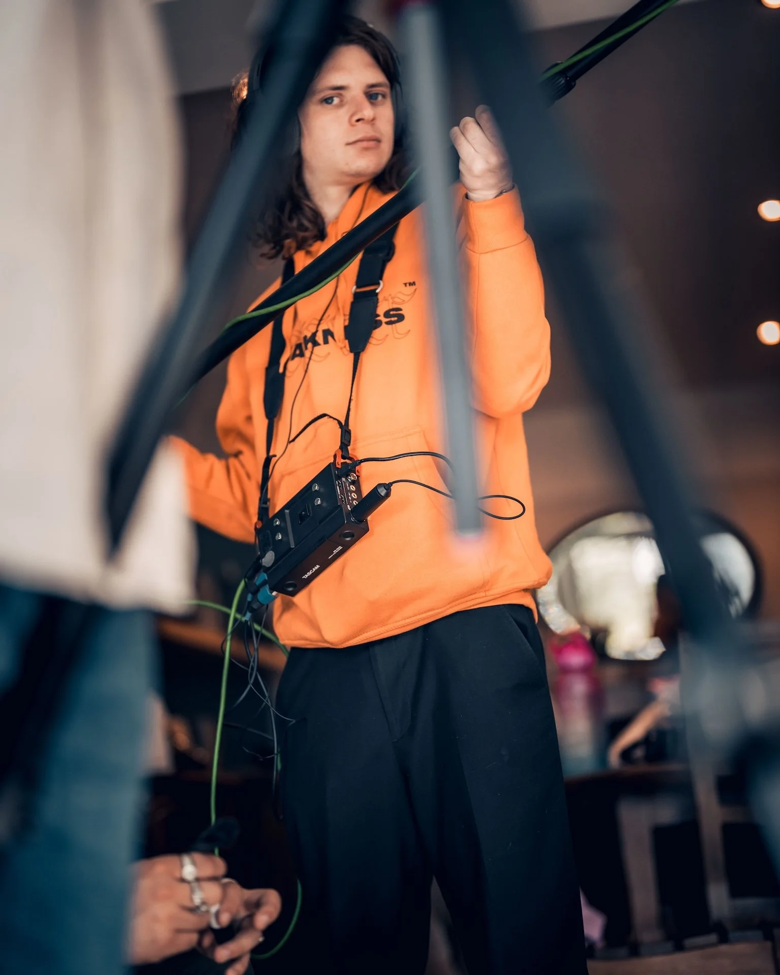 A person with long hair wearing an orange sweatshirt with a logo, using headphones, and operating electronic equipment with cables.