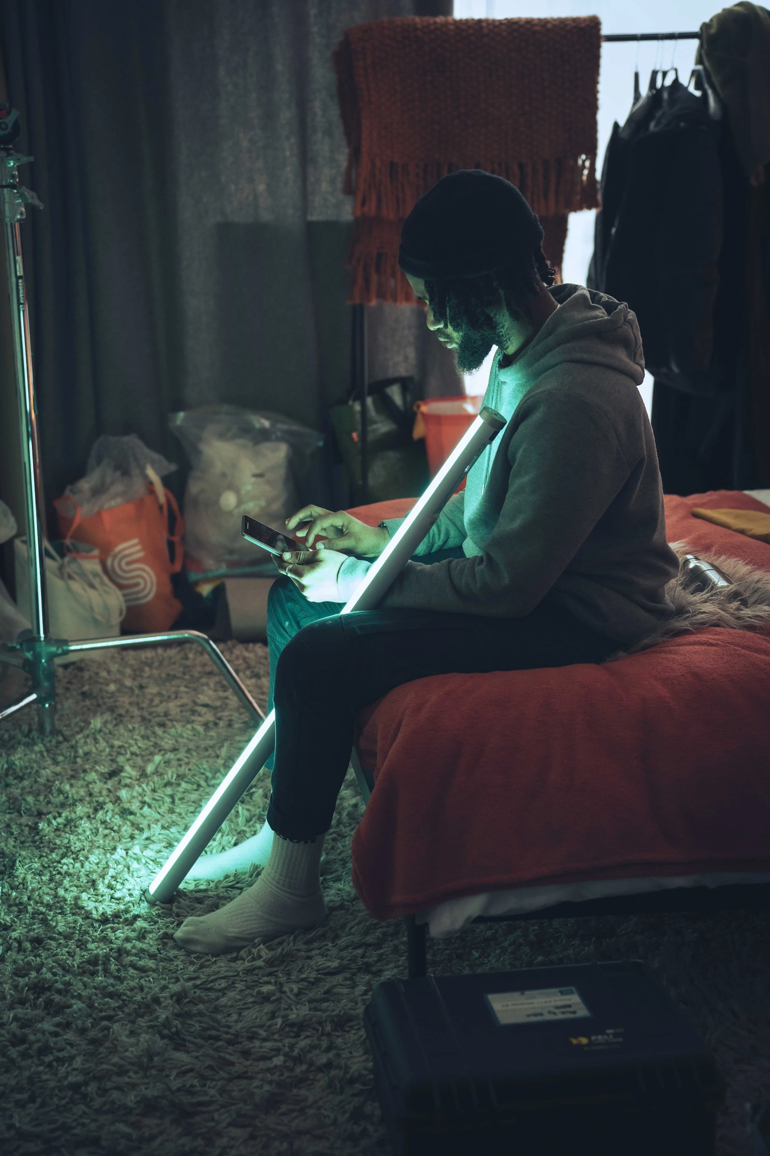 A man with dreadlocks and a beard, wearing a black beanie and hoodie, is sitting on a bed with a orange blanket, looking at his phone. The room is dimly lit with a glow from his phone, with a backdrop of a laundry drying rack, black curtains, and var