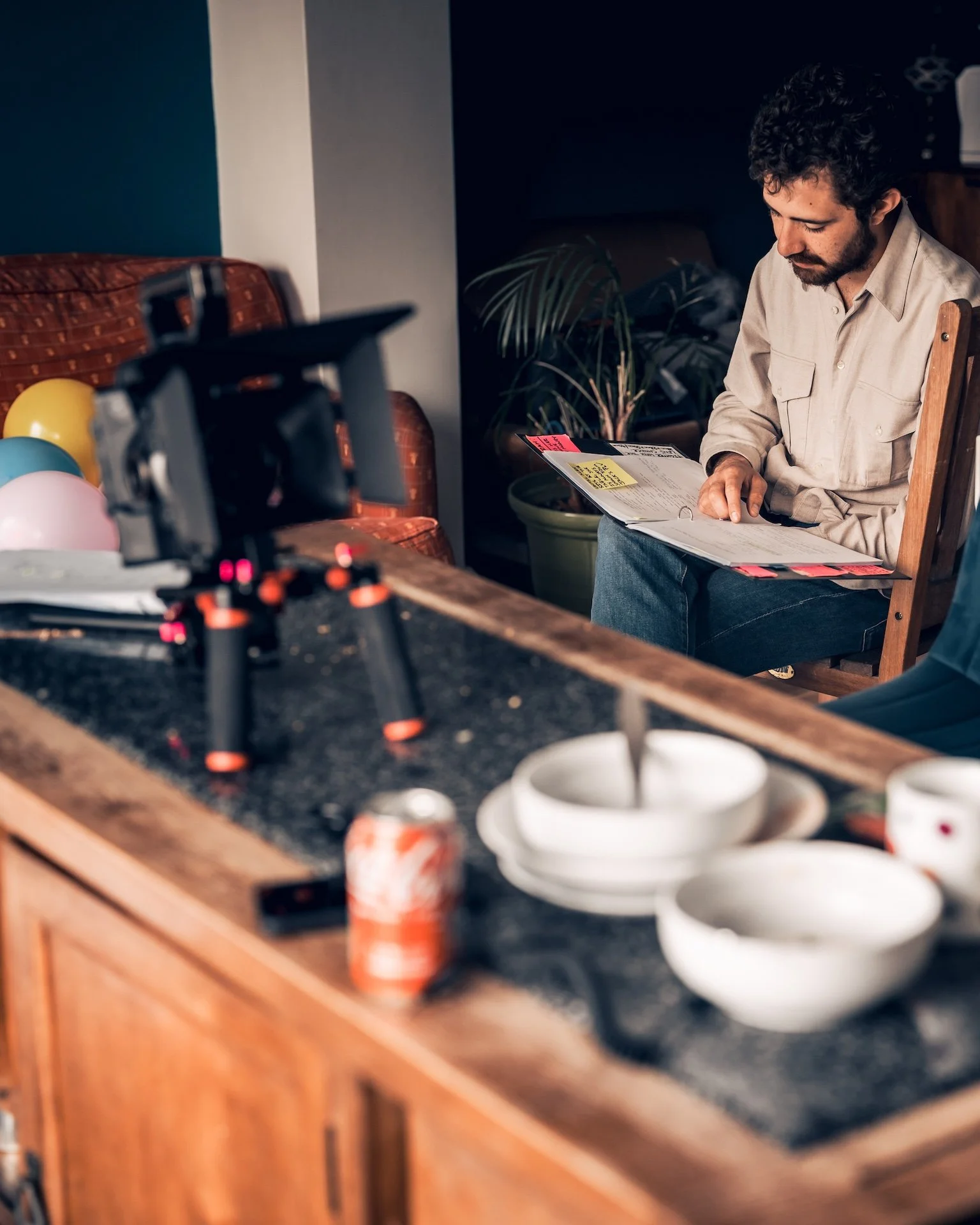 A man with dark curly hair and a beard sitting on a wooden chair in a living room, reading a folder with some colorful sticky notes attached. In the foreground, a kitchen counter with balloons and cups, a toy drone, and soda cans.