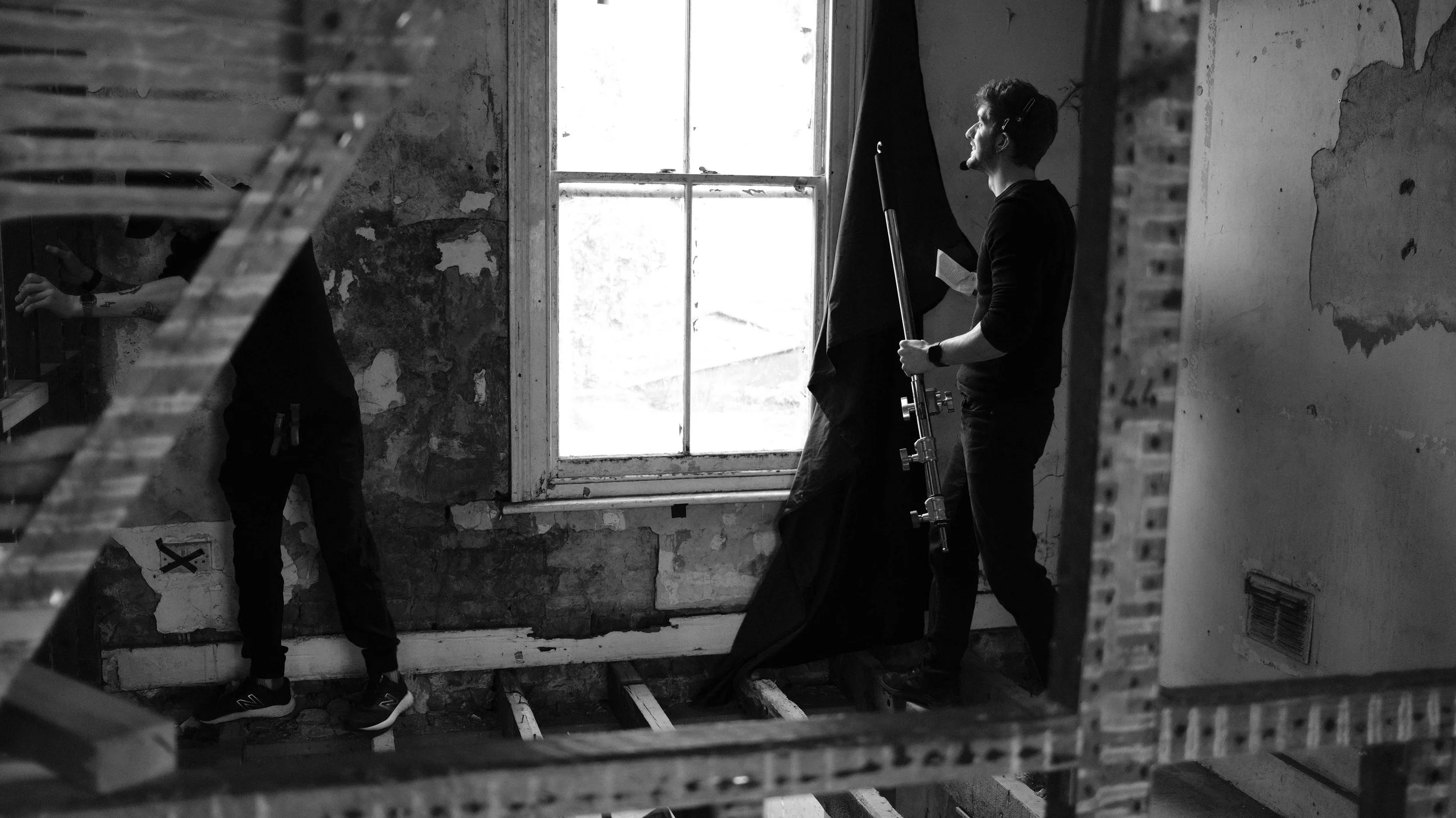 Two men working on a building interior, one adjusting a ladder and the other holding a large black cloth near a window with peeling walls.
