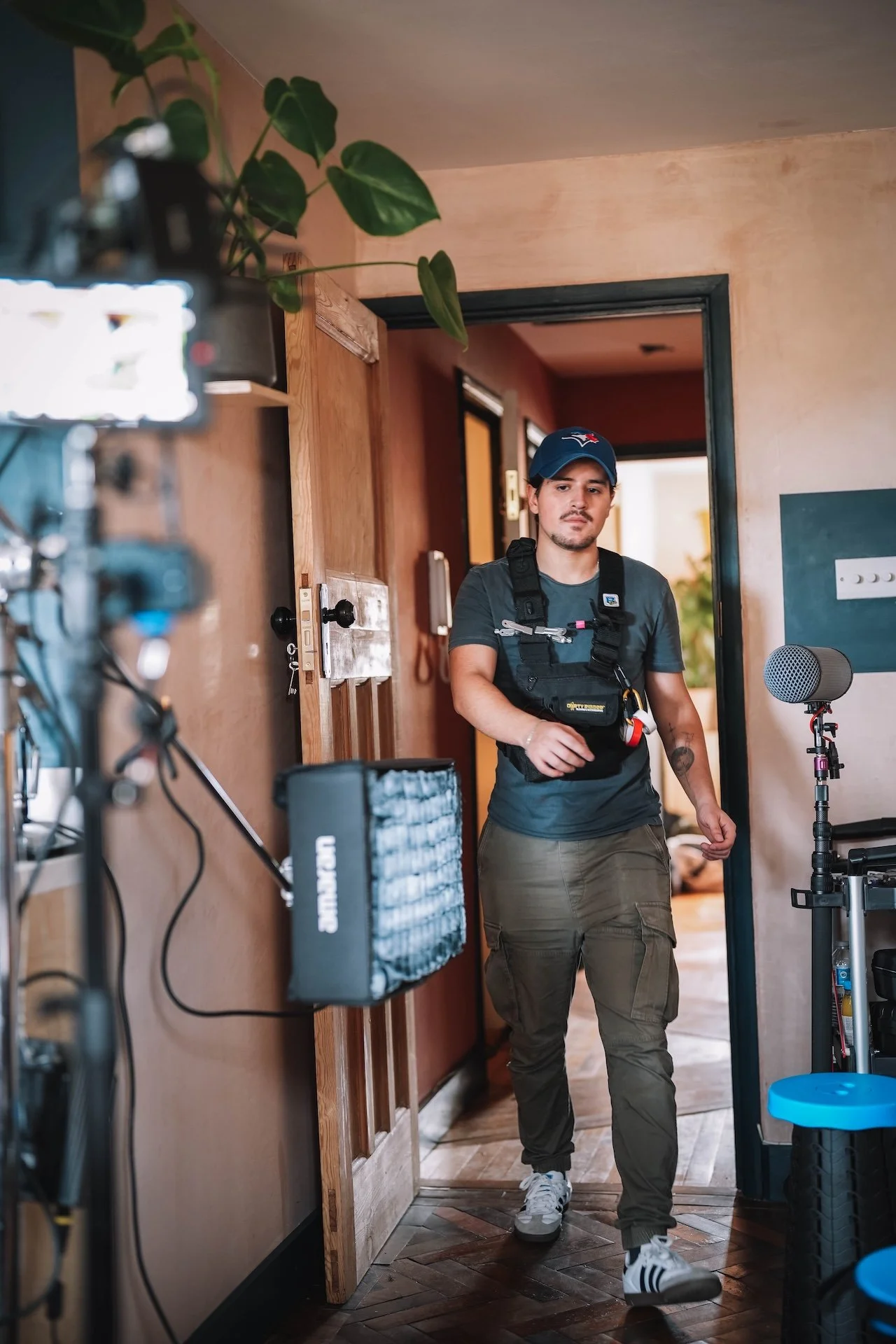 A young man with a goatee, wearing a blue cap with the Toronto Blue Jays logo, a gray T-shirt, cargo pants, and white sneakers, is walking inside a room with a camera set up pointed at him. The room has a wooden doorframe, some equipment, and a potte