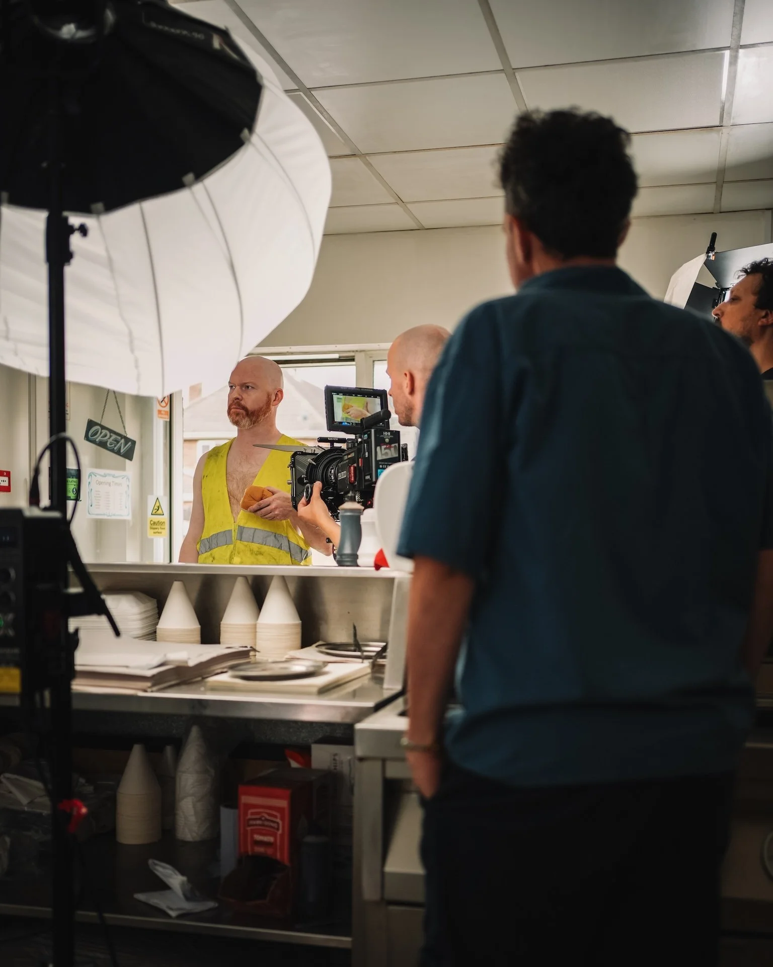 A person with dark curly hair wearing a black shirt stands behind a counter, filming a man in a yellow reflective safety vest with a camera. The man with the vest appears to be talking, with shelves of disposable cups and paper bags behind him. Studi