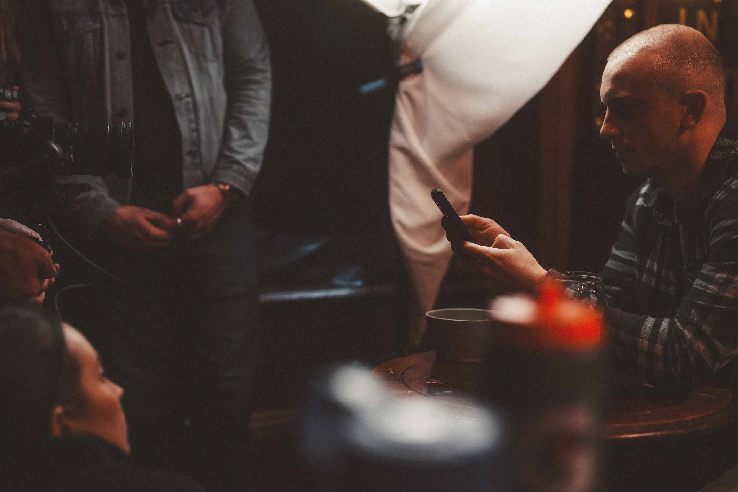 A man with a buzz cut hairstyle sitting at a table in a dimly lit setting examining his phone. Another person standing next to him has hands clasped in front of them. The scene suggests a casual or social environment.