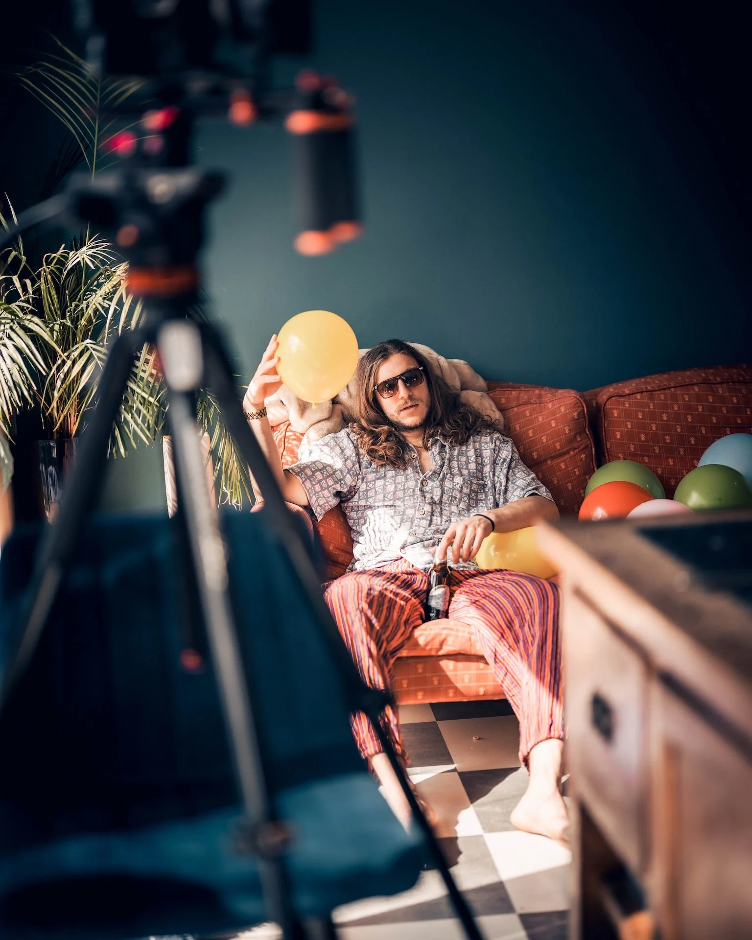 A man with long hair, sunglasses, and casual clothes sitting on a red checkered couch, holding a yellow balloon, surrounded by colorful balloons, with a camera in the foreground and a plant on the left side.