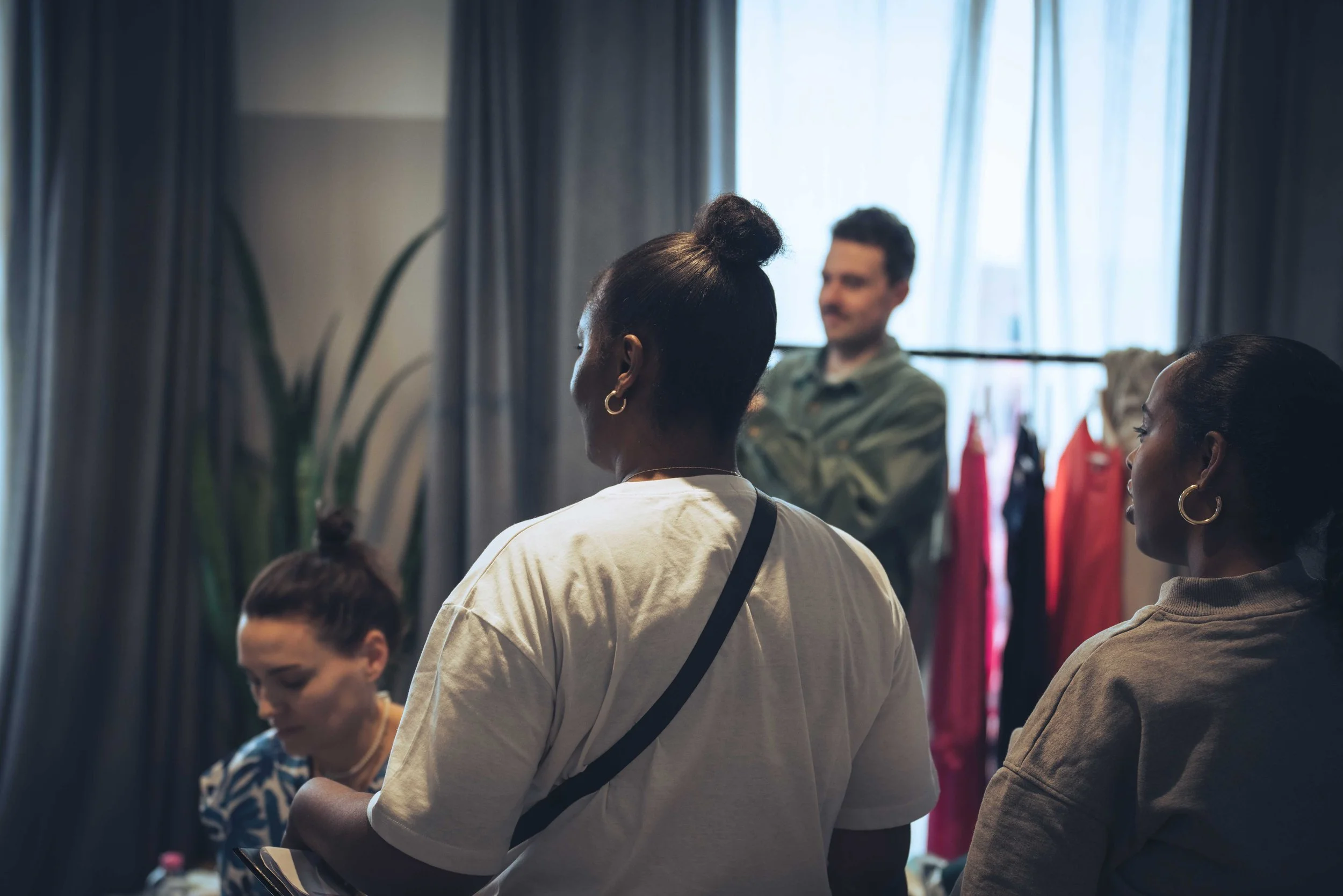 A group of people, including women with their hair styled in buns and others with their hair tied back, are gathered indoors during daylight. They appear to be engaged in a discussion or meeting, with some seated and some standing. The background fea