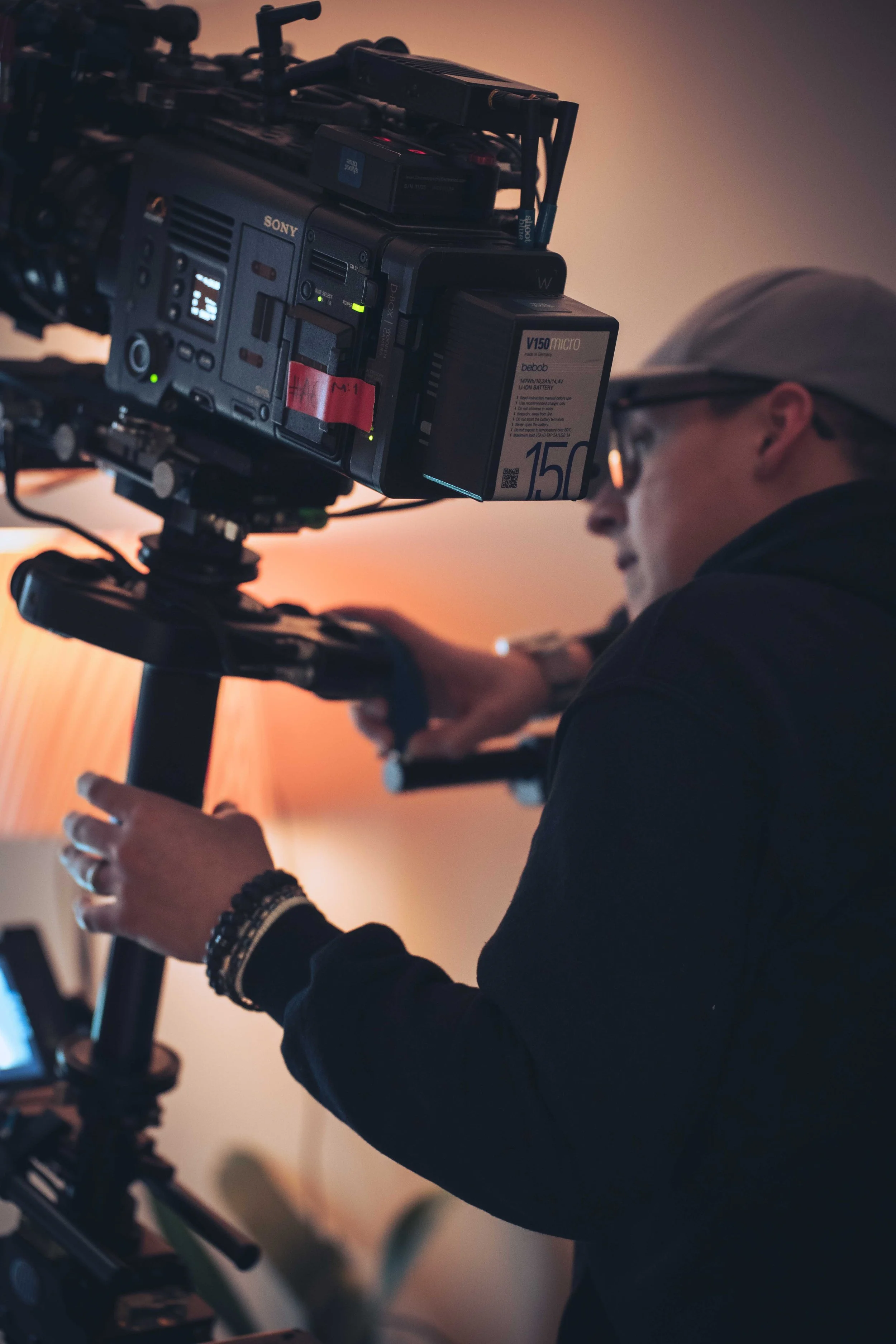 A person wearing a light-colored cap and glasses operating a professional Sony video camera on a tripod, with a sunset sky in the background.