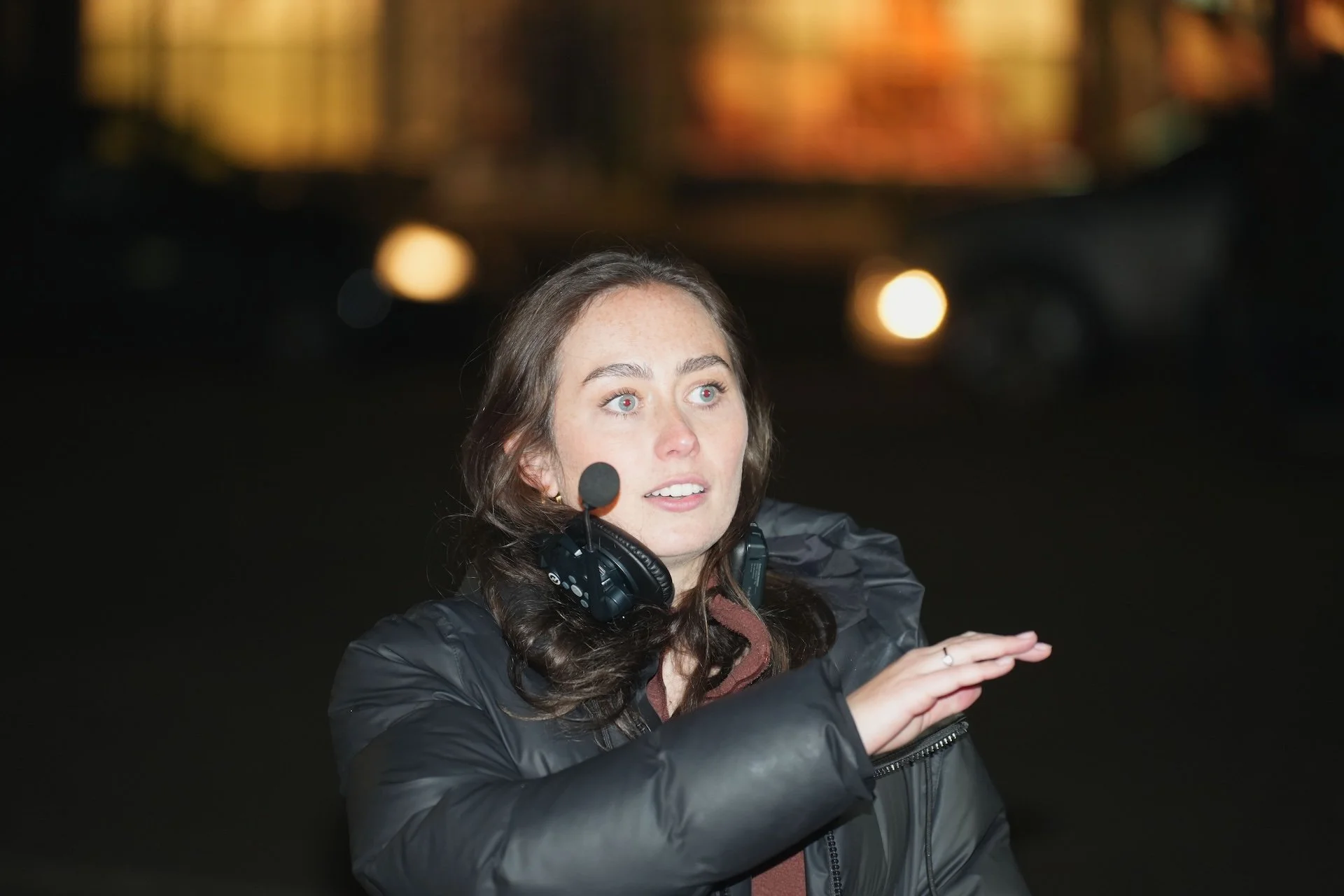 A woman with long brown hair, wearing a black jacket and a headset with a microphone, speaking or gesturing with her hands in an indoor setting with blurry background lights.