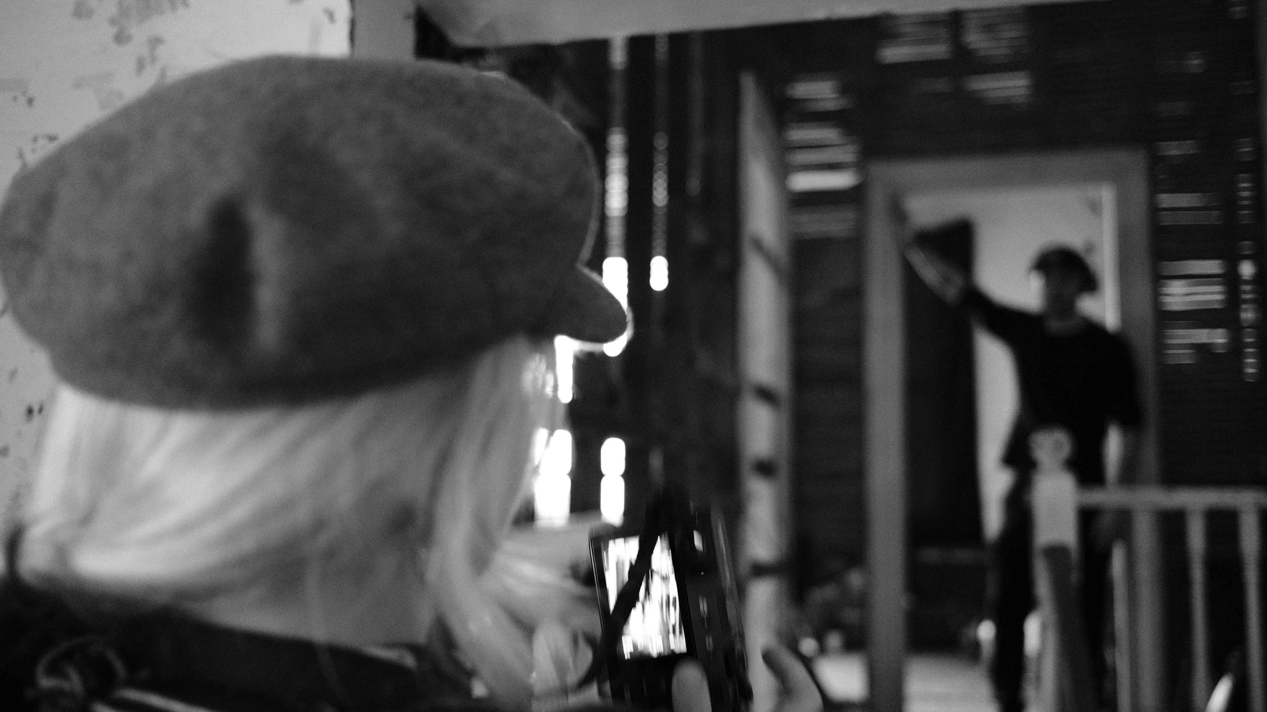 A person with a hat taking a photo or video of another person holding a hat, cloaked in shadow, on a wooden porch or deck.