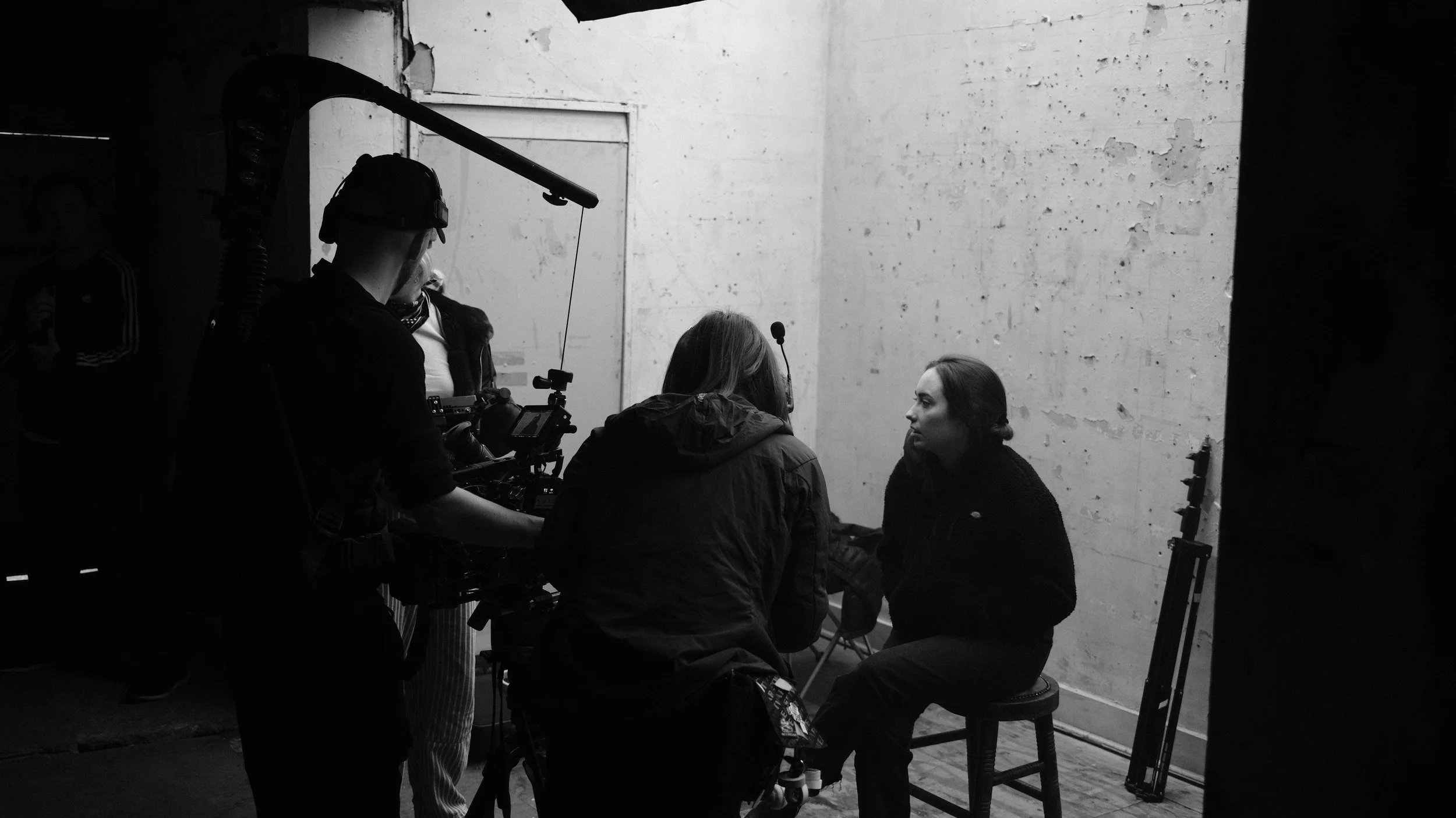 Black and white photo of a film crew filming a woman sitting on a stool in front of a plain wall.