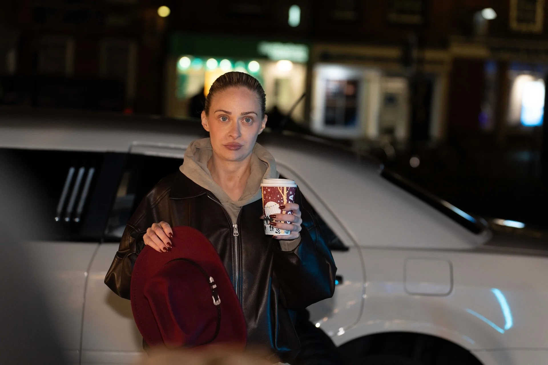 A woman with dark hair tied back, wearing a black leather jacket and gray hoodie, holding a holiday-themed cup in her right hand and a red hat in her left hand, standing next to a white car on a city street at night.
