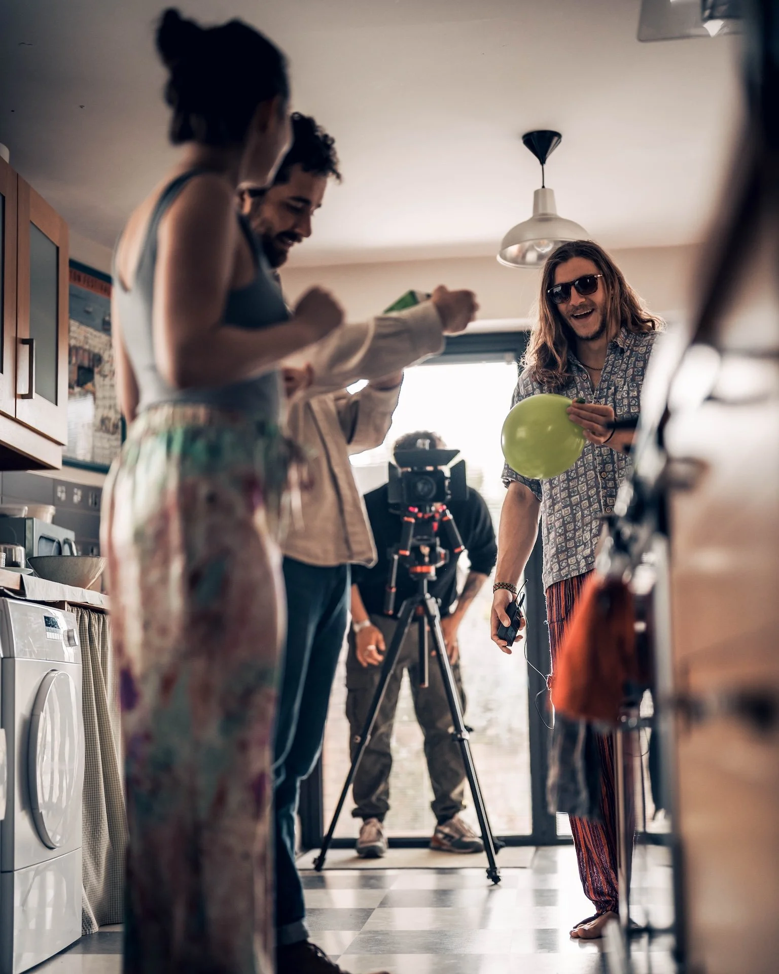 Three men in a kitchen, one holding green balloon, another operating camera on tripod, a woman in background, all smiling.
