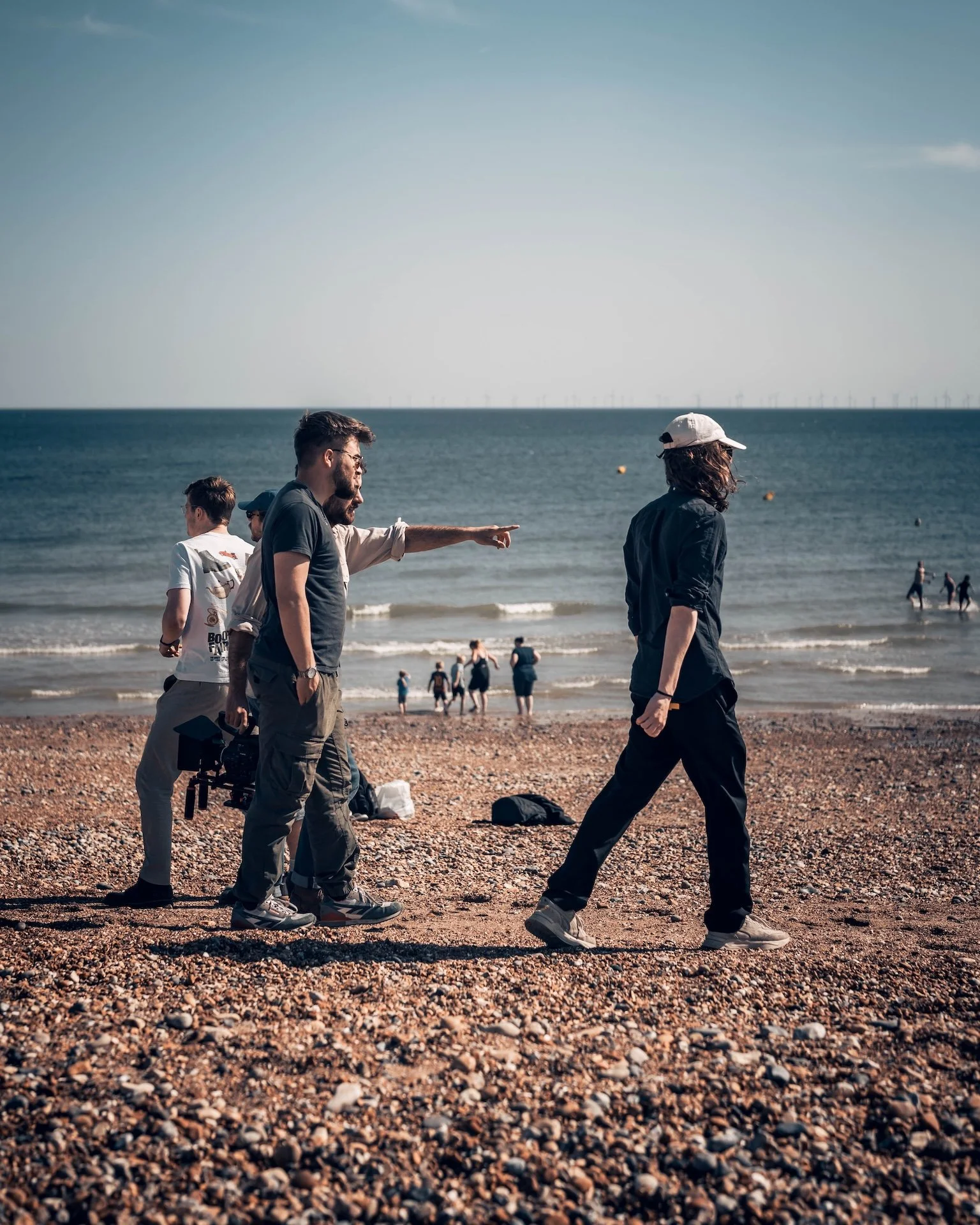 People walking on a pebble beach near the ocean, with some engaging in conversation and others in the background near the water.