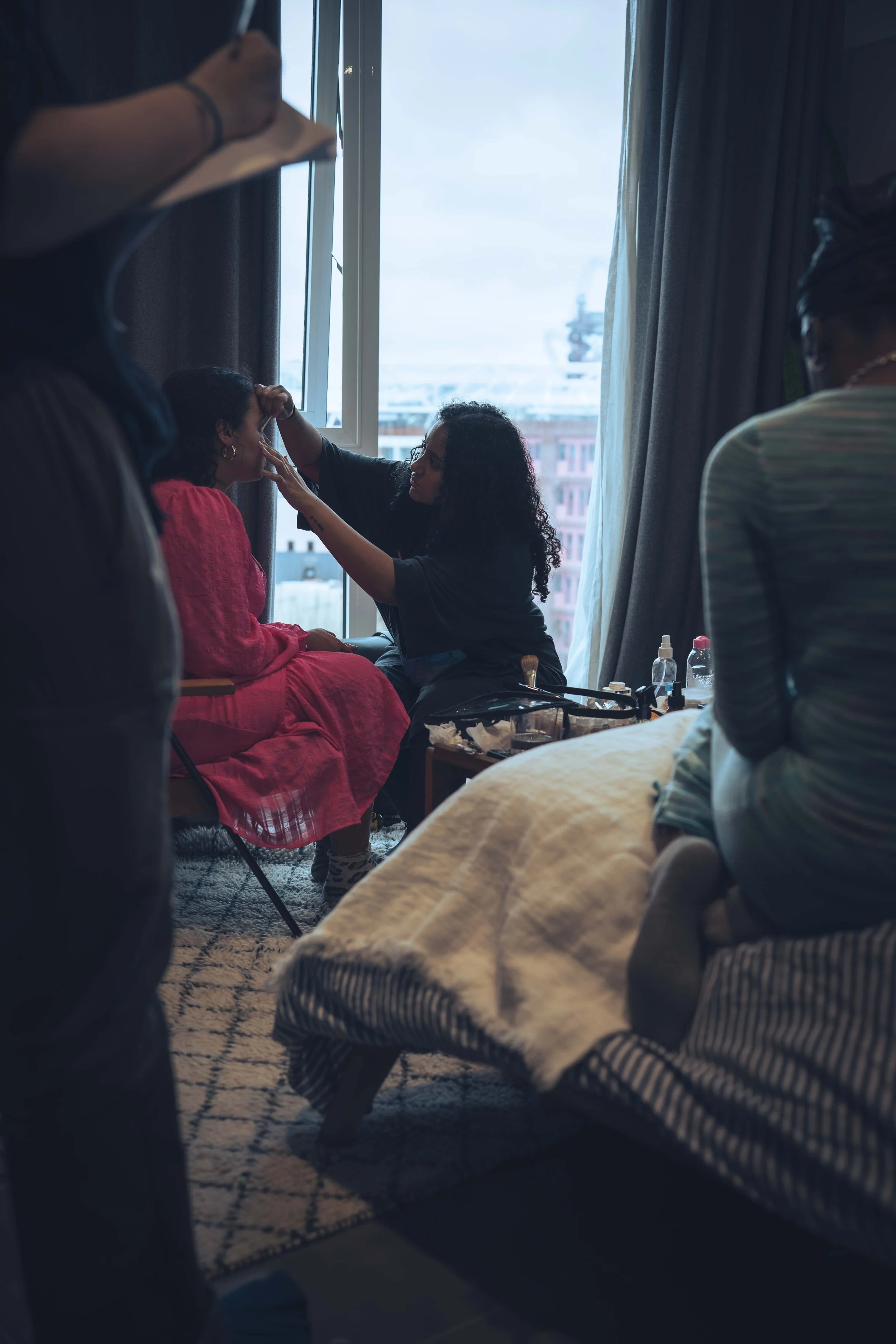 A woman is applying makeup to a girl seated by a window in a room. There are two other women in the room, one near the camera and another on the right side, with makeup supplies on a table. The room has a patterned rug and a window with curtains, sho