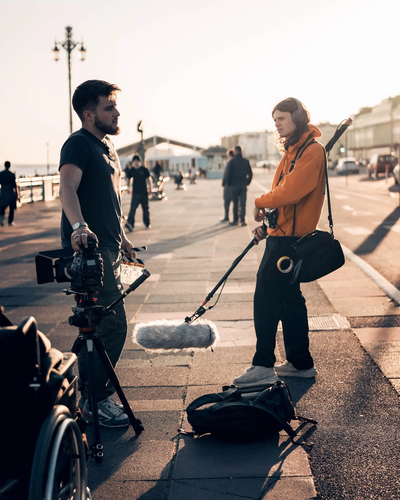 Two men engaged in conversation on a boardwalk during sunset. One is holding a camera and film slate, the other has headphones and carries filming equipment. People and cars are visible in the background.