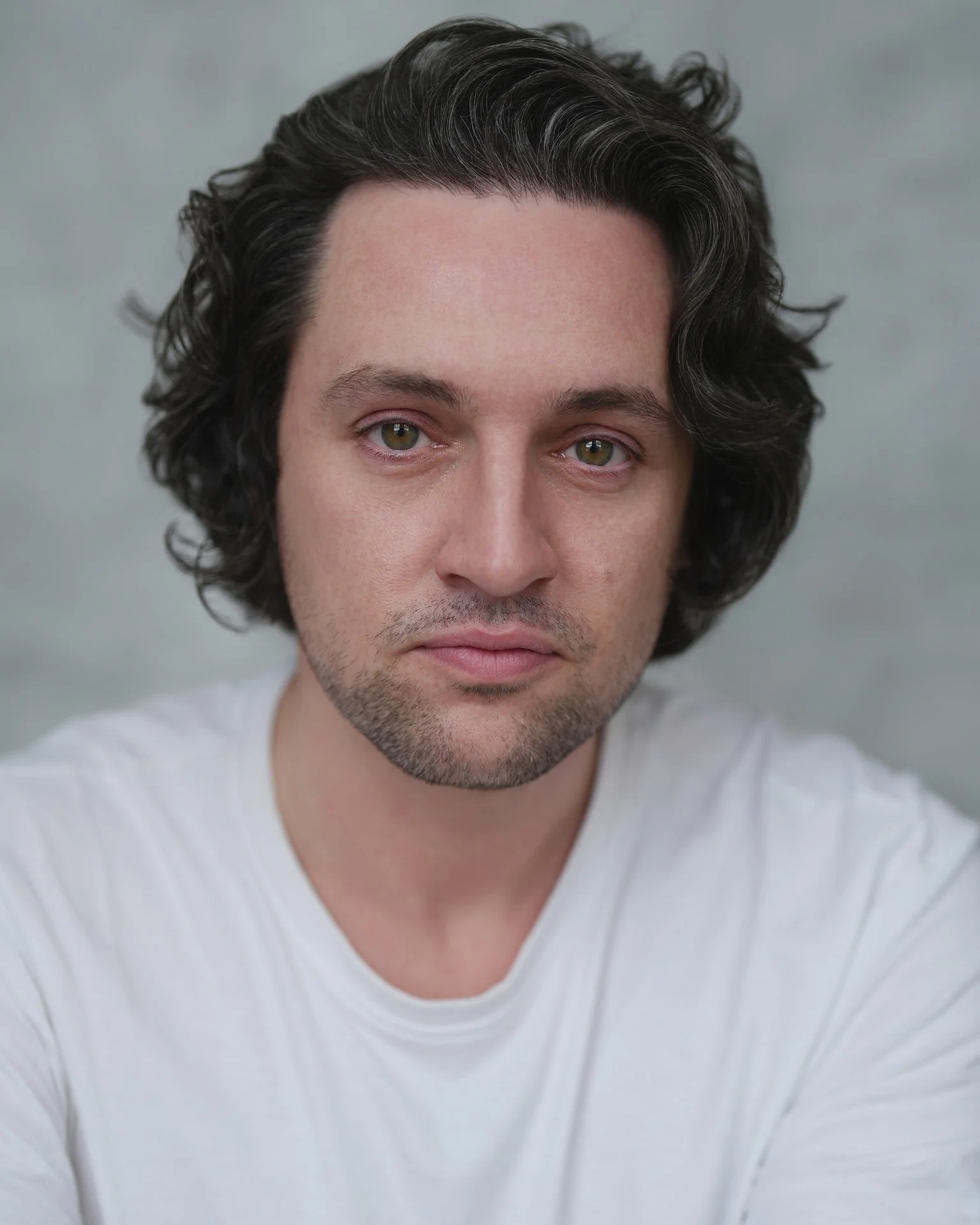 Close-up portrait of a man with wavy dark hair, light skin, and wearing a white T-shirt, looking directly at the camera.