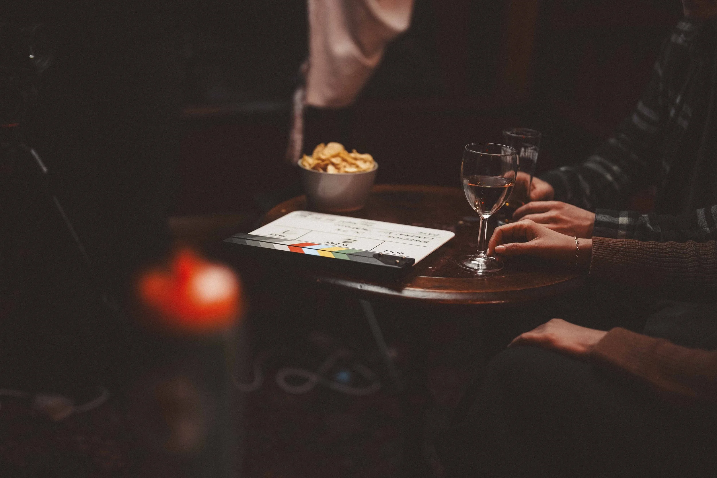 A film slate, a glass of pink wine, a bowl of potato chips, and a person's hand with a bracelet are on a wooden table in a dimly lit setting.