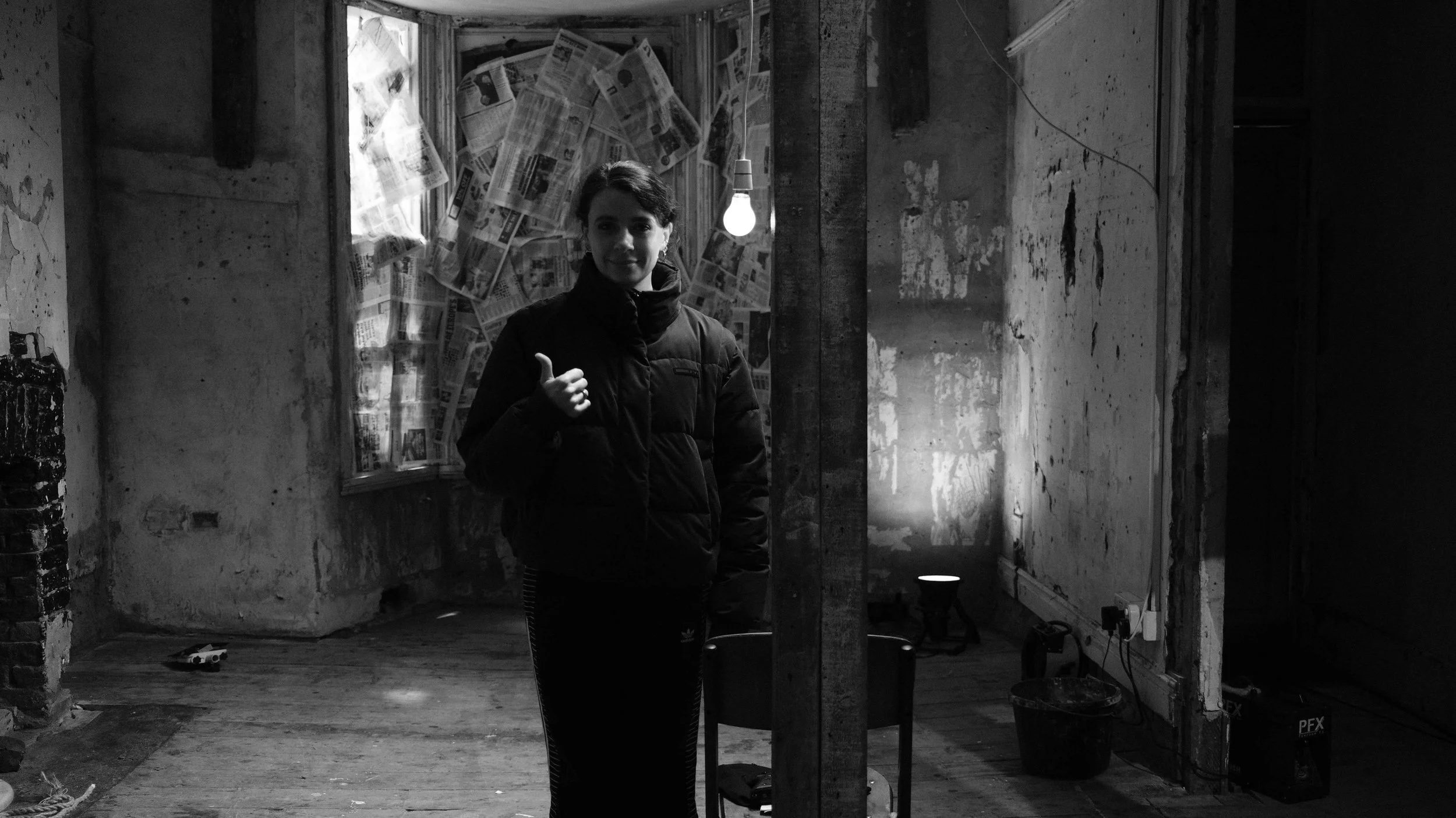 A woman in a puffy jacket giving a thumbs up inside a rustic, partially unfinished room with exposed brick, wooden beams, paper-covered window, and visible wiring.