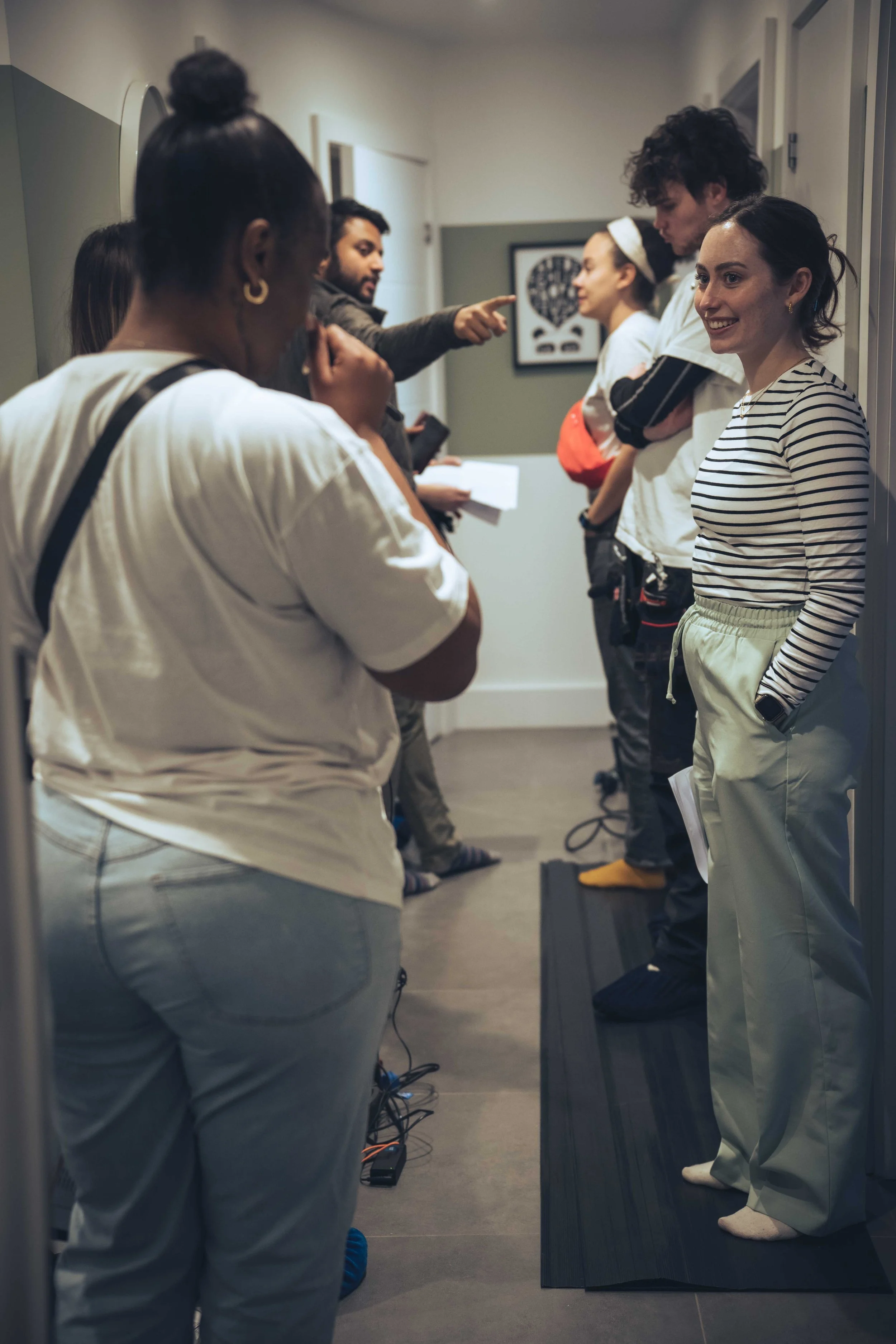 A group of people waiting in a hallway. Some are standing with crossed arms, some are looking at their phones or talking. The floor is dark hardwood, and a picture is hanging on the wall in the background.