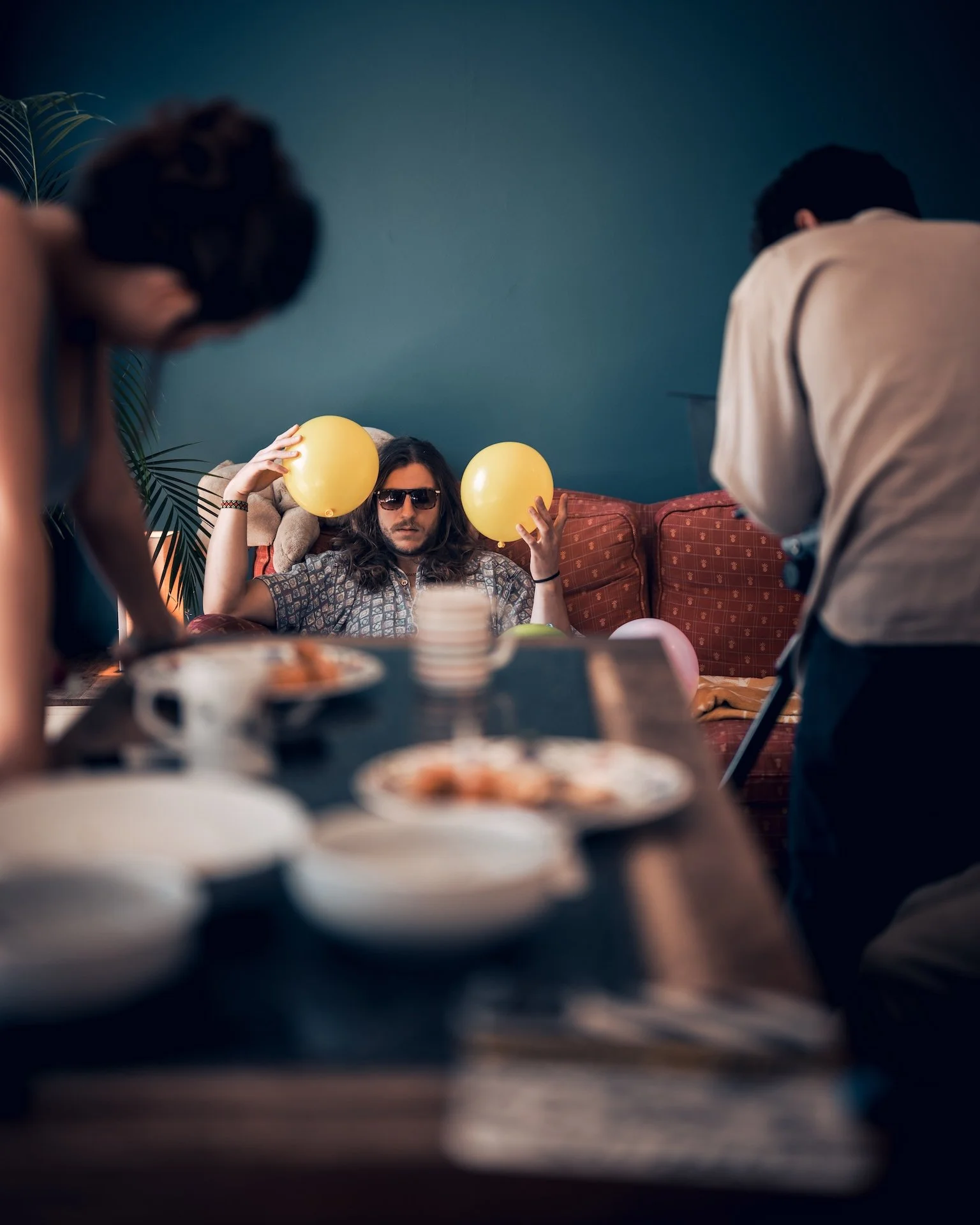 A person with long hair wearing sunglasses holding two yellow balloons, sitting on a sofa with a teal wall background, surrounded by party decorations and snacks on a table in the foreground.