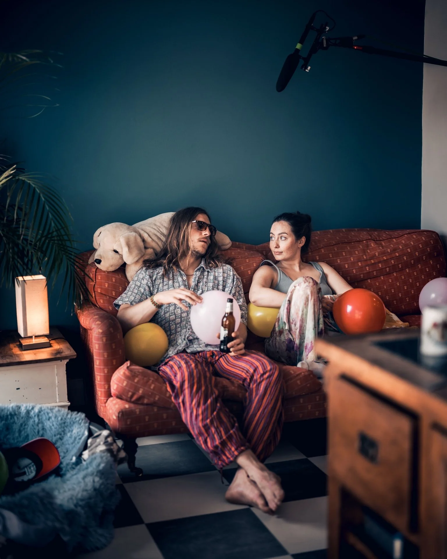 Two young women sitting on a red sofa in a room with a blue wall, surrounded by balloons, party decor, and scattered items, with a microphone hanging overhead, indicating a casual social gathering.