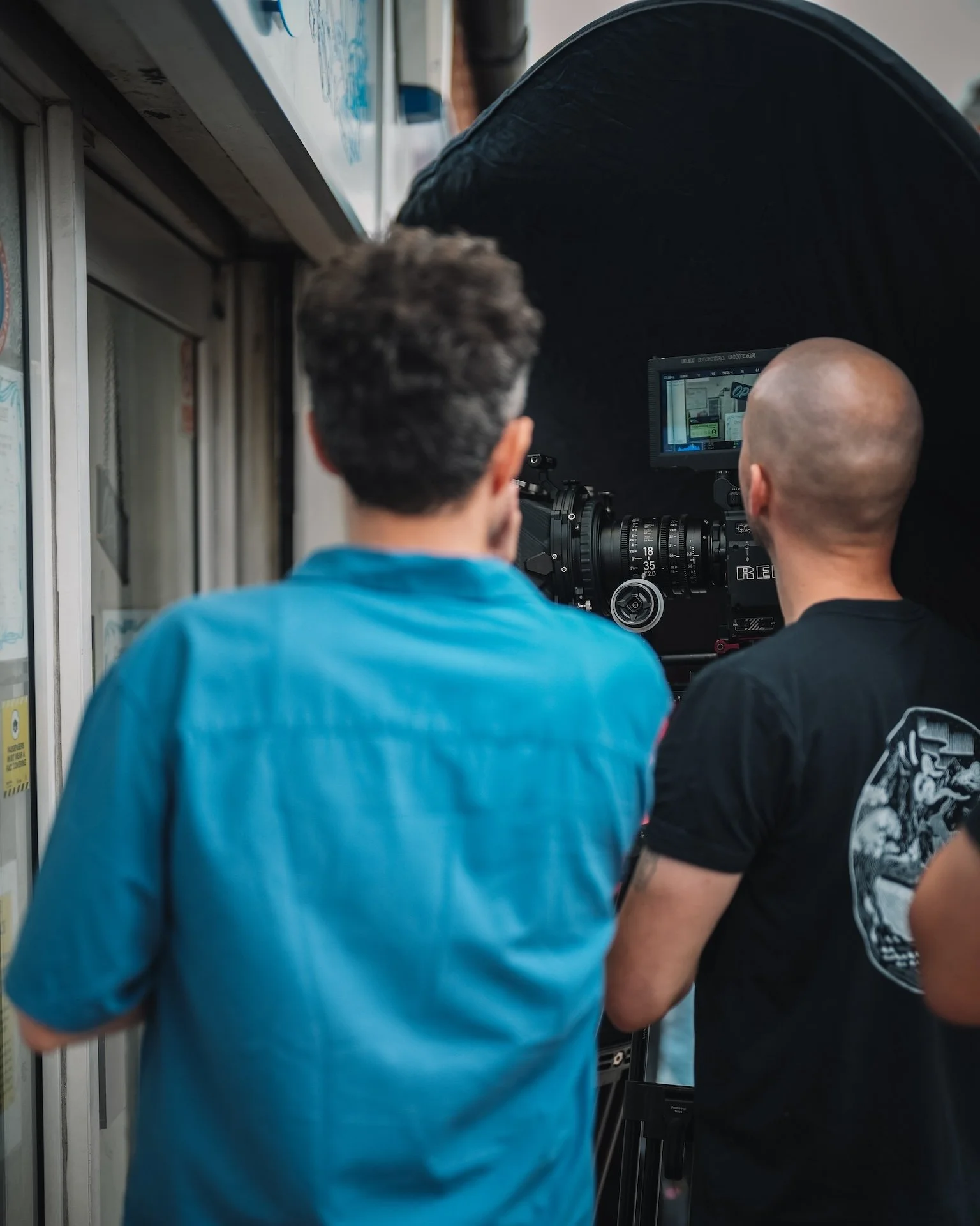 Two men working together with a professional camera on a tripod, filming or photographing at an outdoor location, possibly near a building with a glass door.