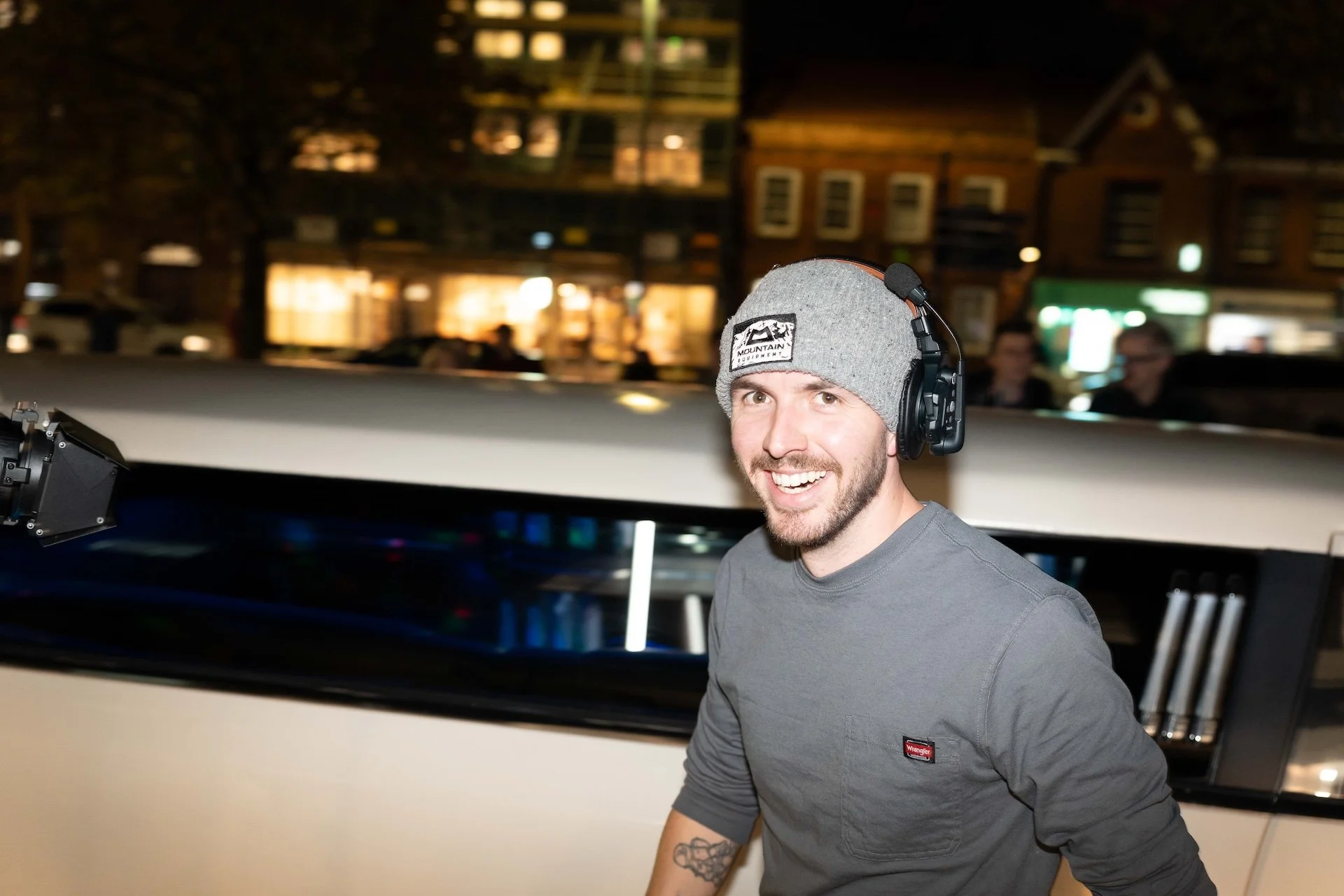 A smiling man wearing a gray beanie, headphones, and a gray shirt standing in front of a white vehicle at night with buildings and blurred lights in the background.
