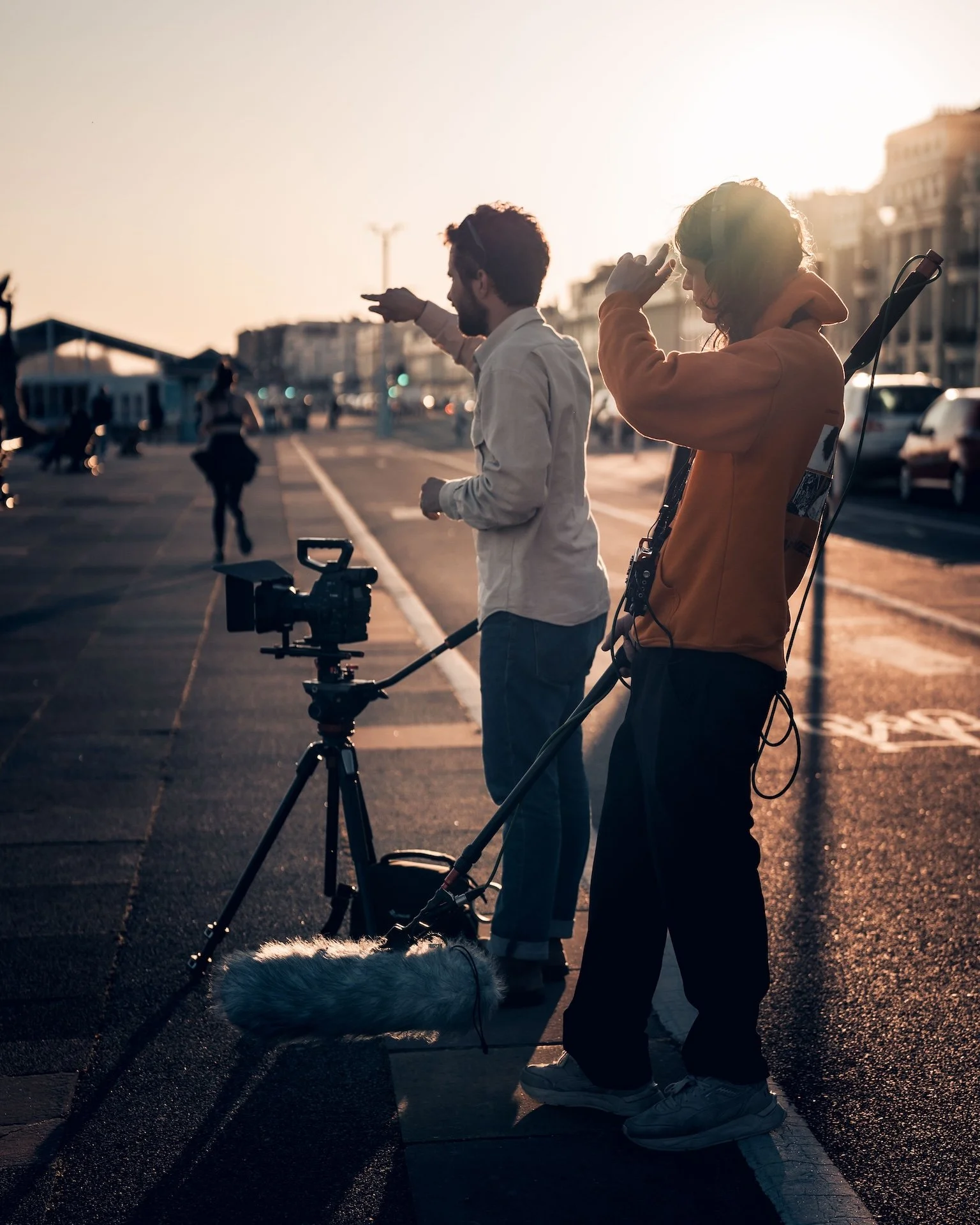 Filmmakers or videographers working outdoors at sunset with a camera on a tripod, a boom microphone, and a woman adjusting her hair. Several people can be seen in the background.