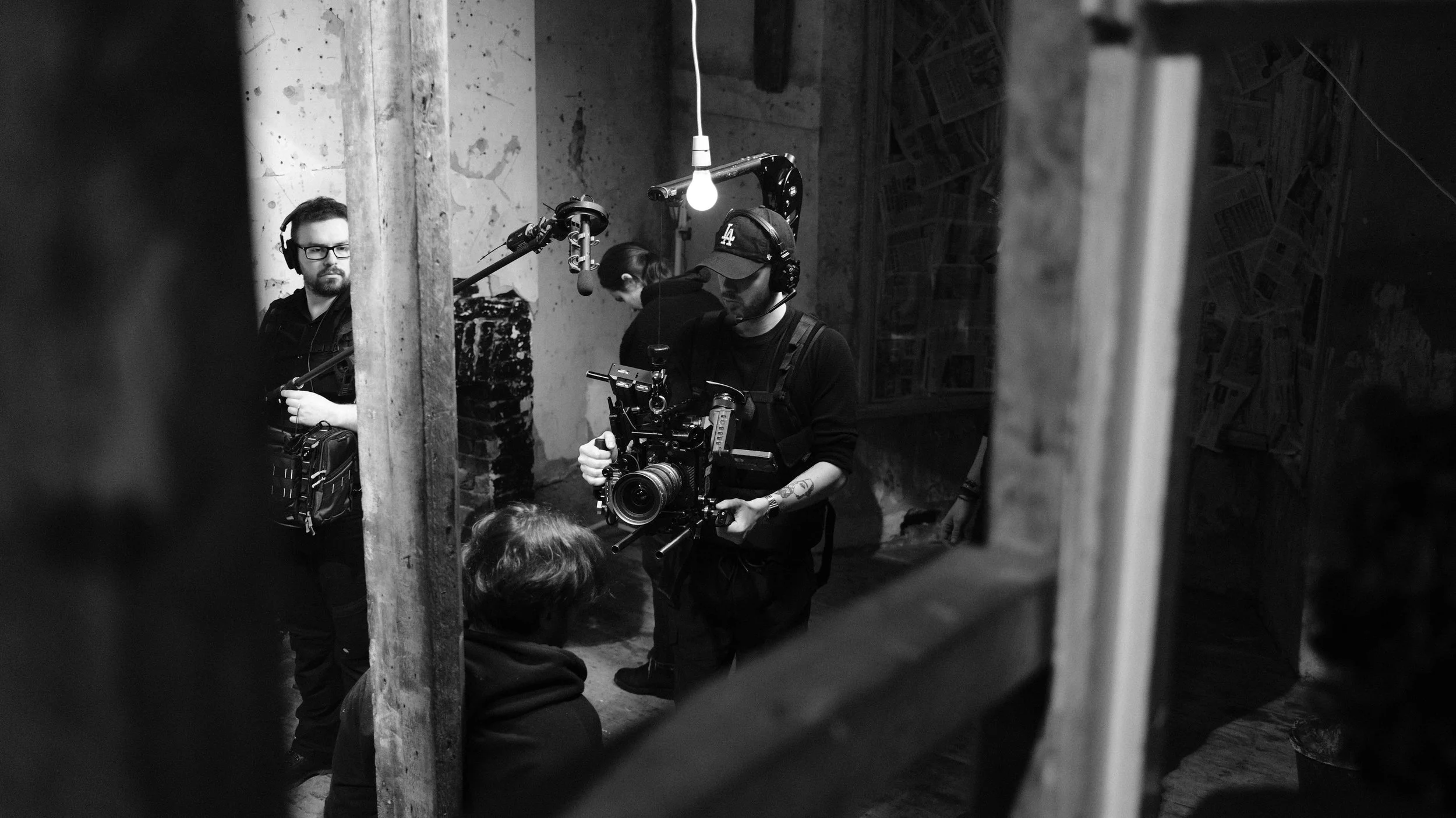 Filmmakers and crew working on a film set, with a camera operator holding a professional camera and crew members wearing headphones, in a room with rough walls and minimal lighting.