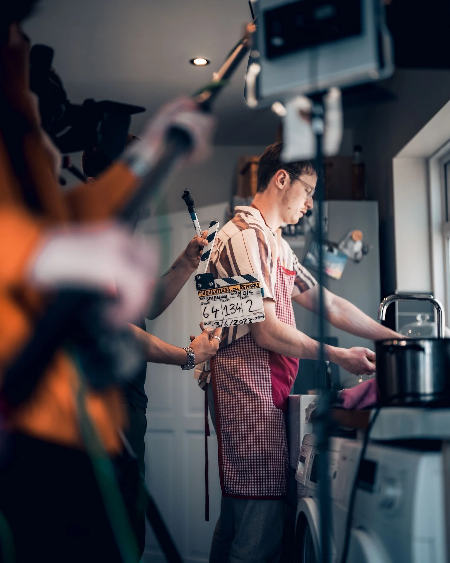 A man in a red checkered apron doing laundry with a washing machine, while a person nearby holds a film clapperboard.