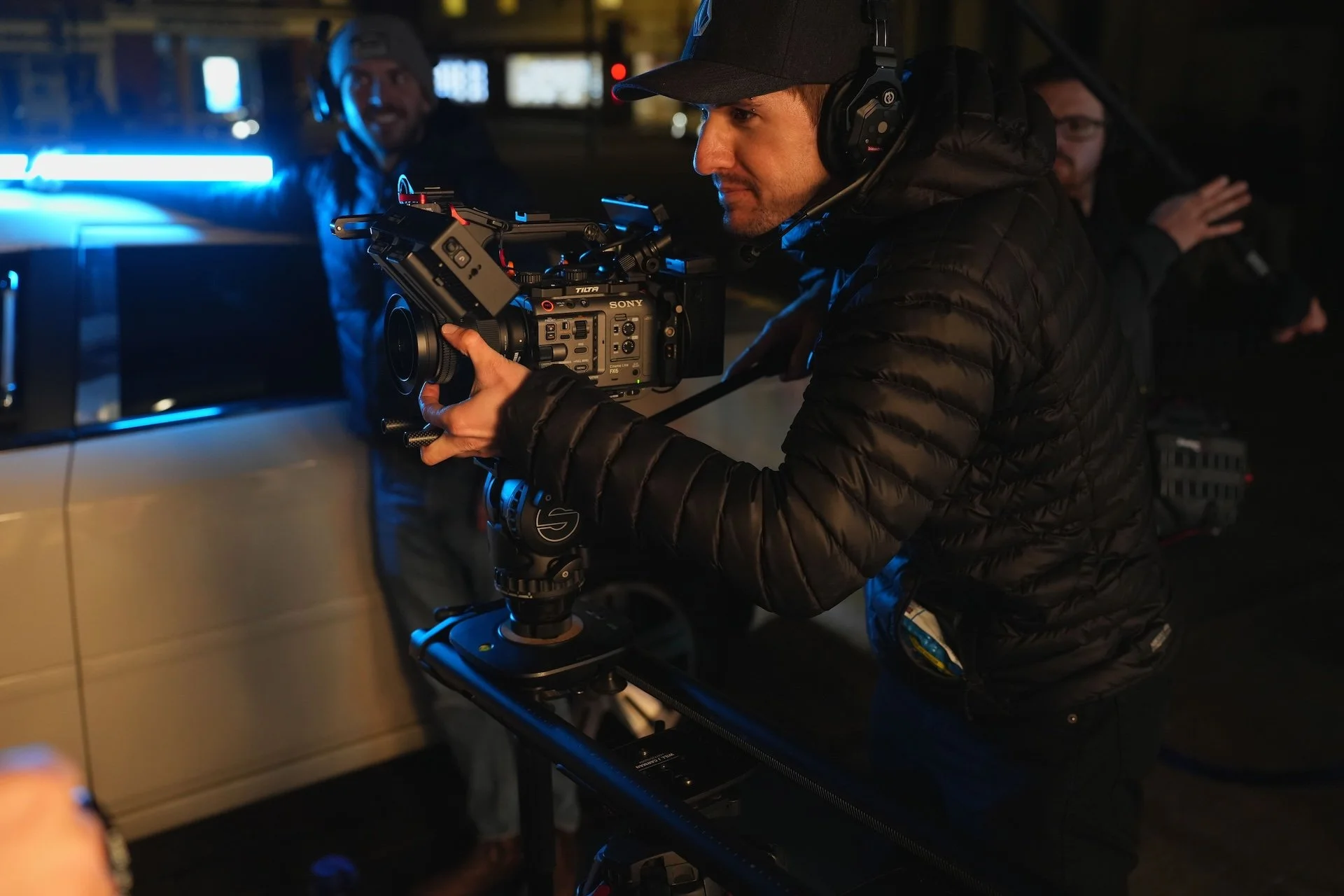A man wearing a black cap and jacket is operating a professional film camera with a Sony logo, while two other crew members are visible in the background in a dimly lit setting.