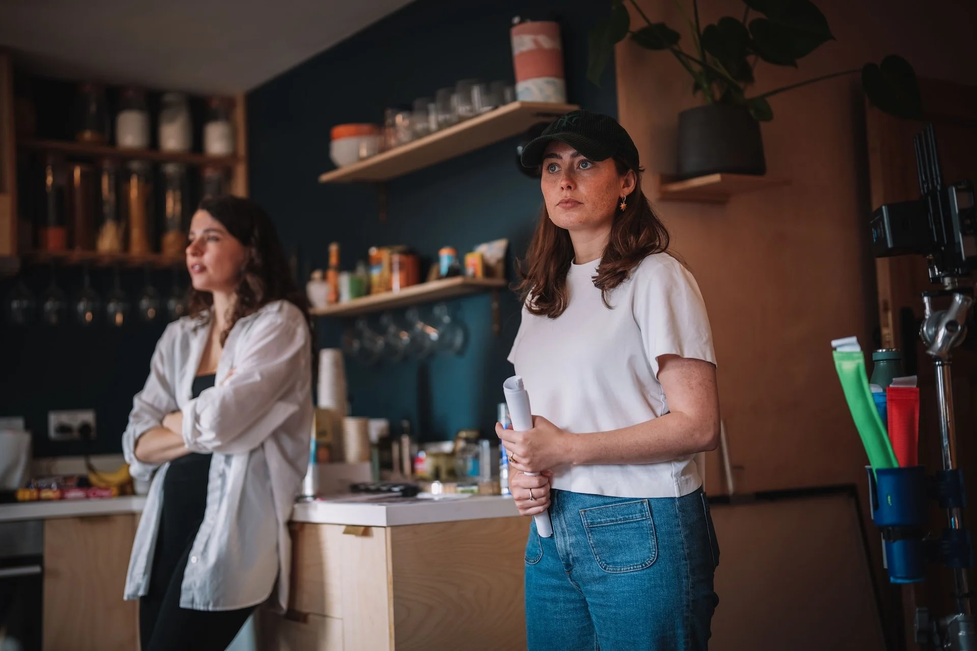 Two women in a kitchen, one holding papers, with shelves of jars and glasses in the background.