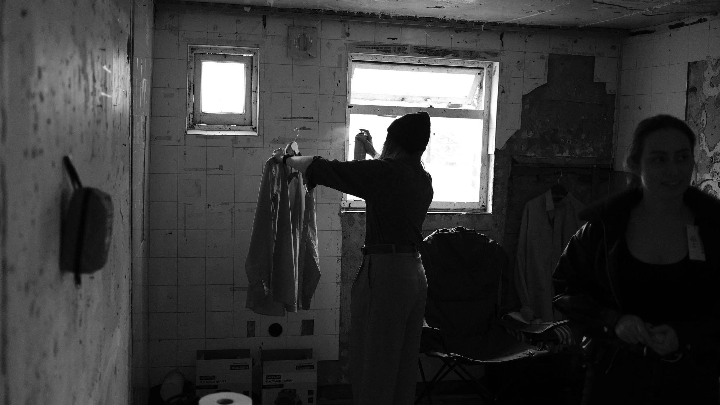 Two women in a dark, dilapidated room with peeling walls, one hanging laundry and the other smiling.