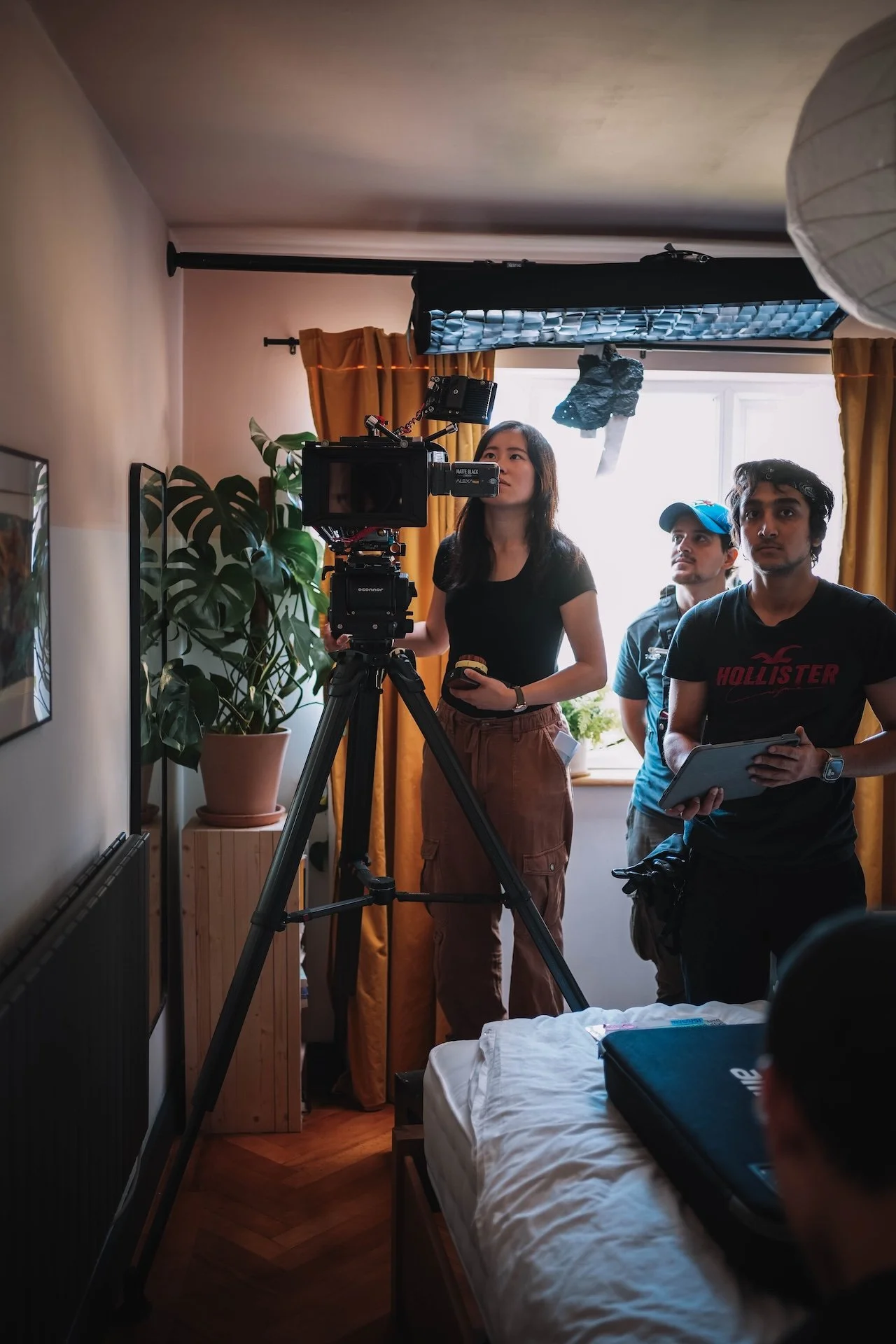 Three people on a film set inside a bedroom with yellow curtains and a large plant, one person is operating a camera, another holds a tablet, and a third is standing behind them.