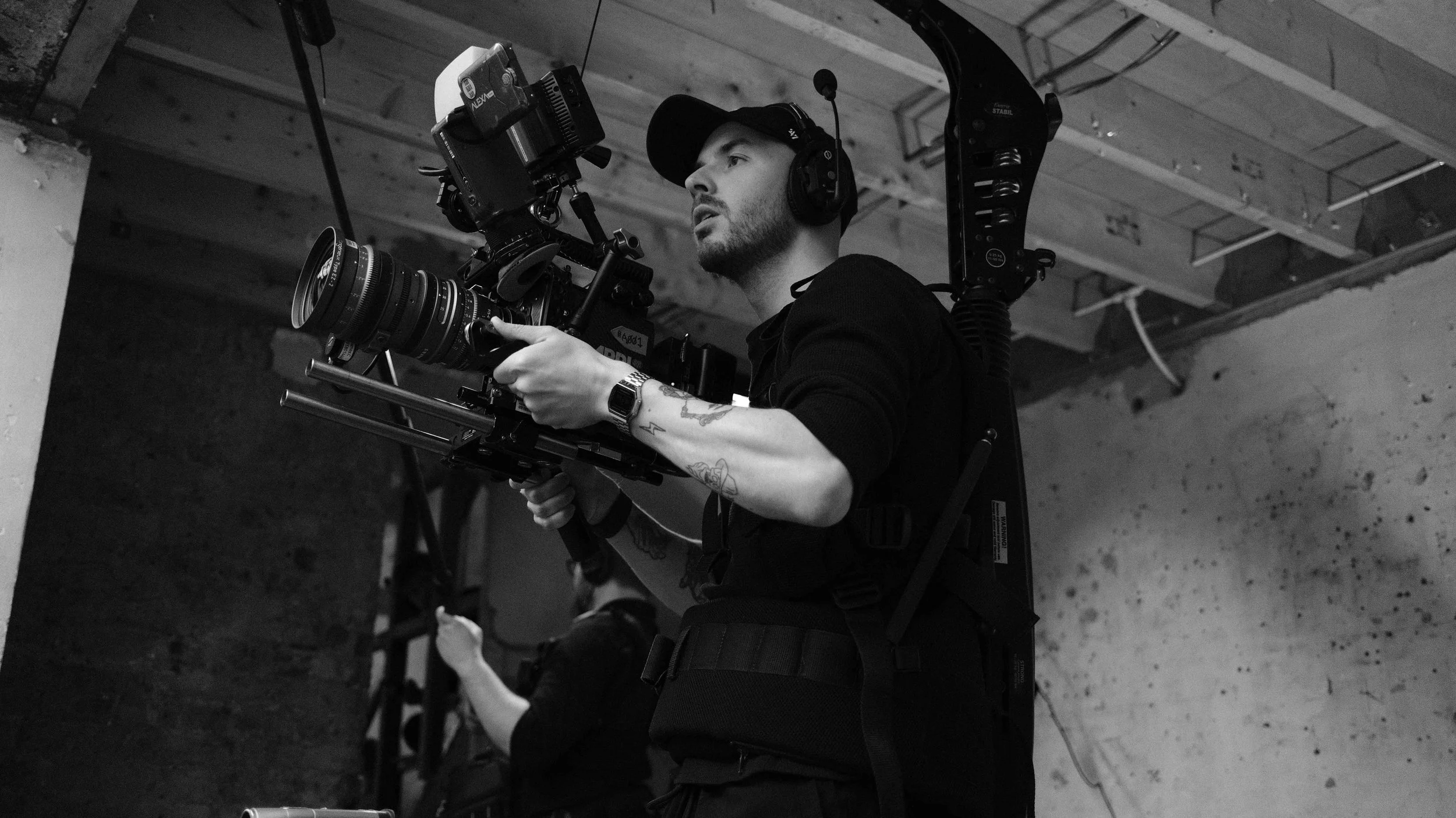 A man operating a professional film camera on a rig, wearing headphones and a cap, with a tattooed arm, in an industrial setting.