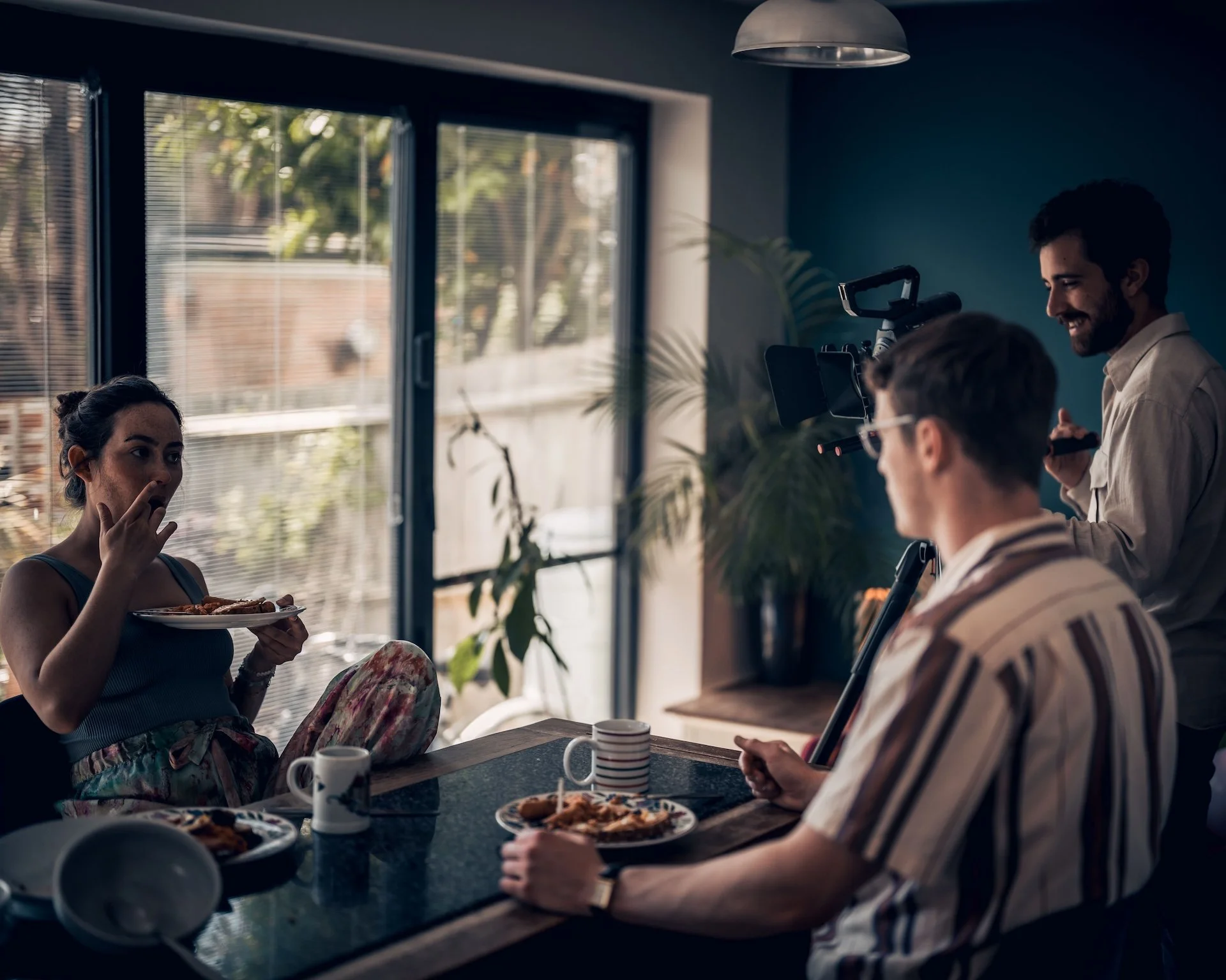 People gathered around a table inside a room with large windows, enjoying pizza and coffee, with a woman on the left about to eat pizza and smiling man on the right talking, camera equipment visible in the background.