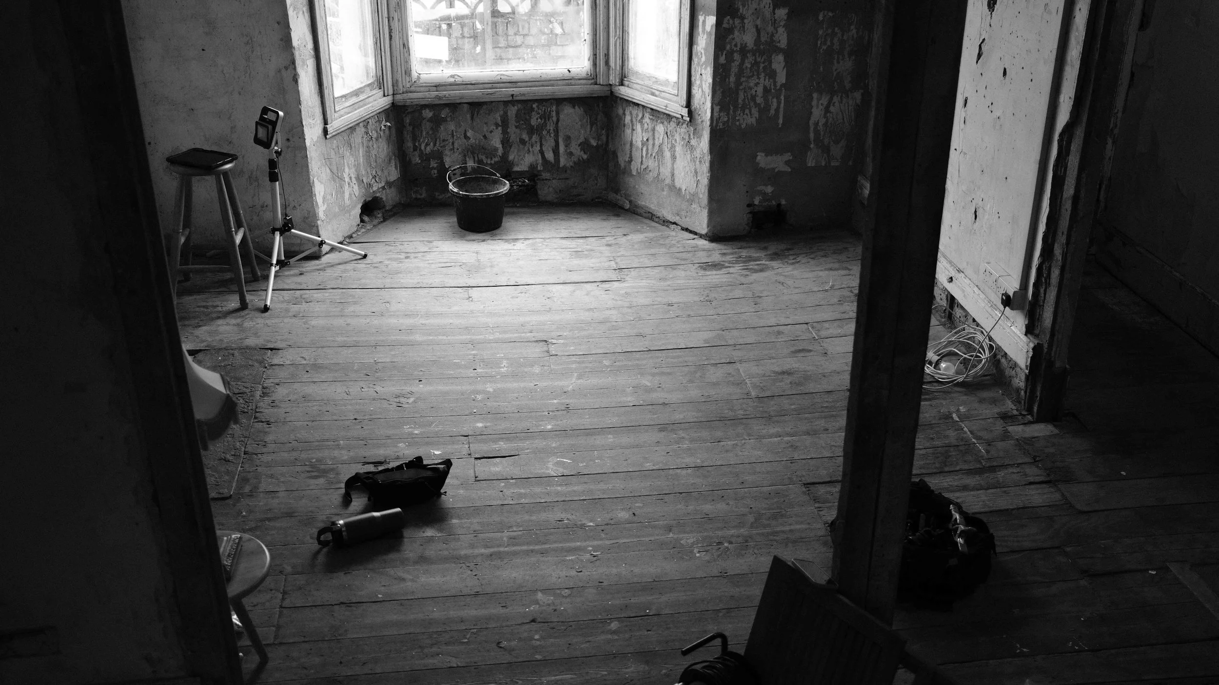 Empty room with unfinished walls and wooden floor, a stool with a tripod and camera, a bucket, a water bottle, a small toolbox, and some scattered cables and tools, with a large window letting in light.