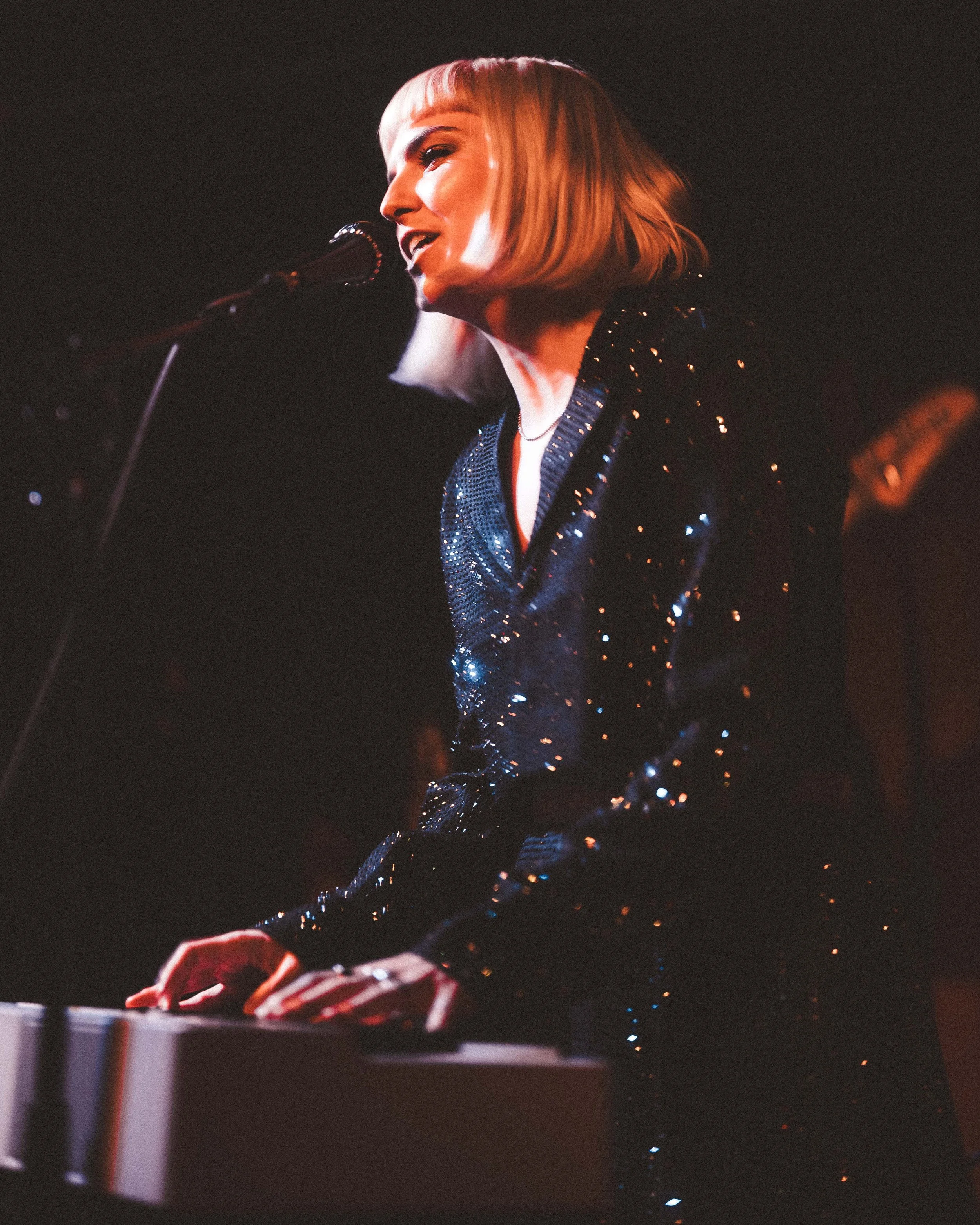 A woman with blonde hair in a bob haircut, wearing a black sequined jacket, playing a keyboard on stage under warm lighting.