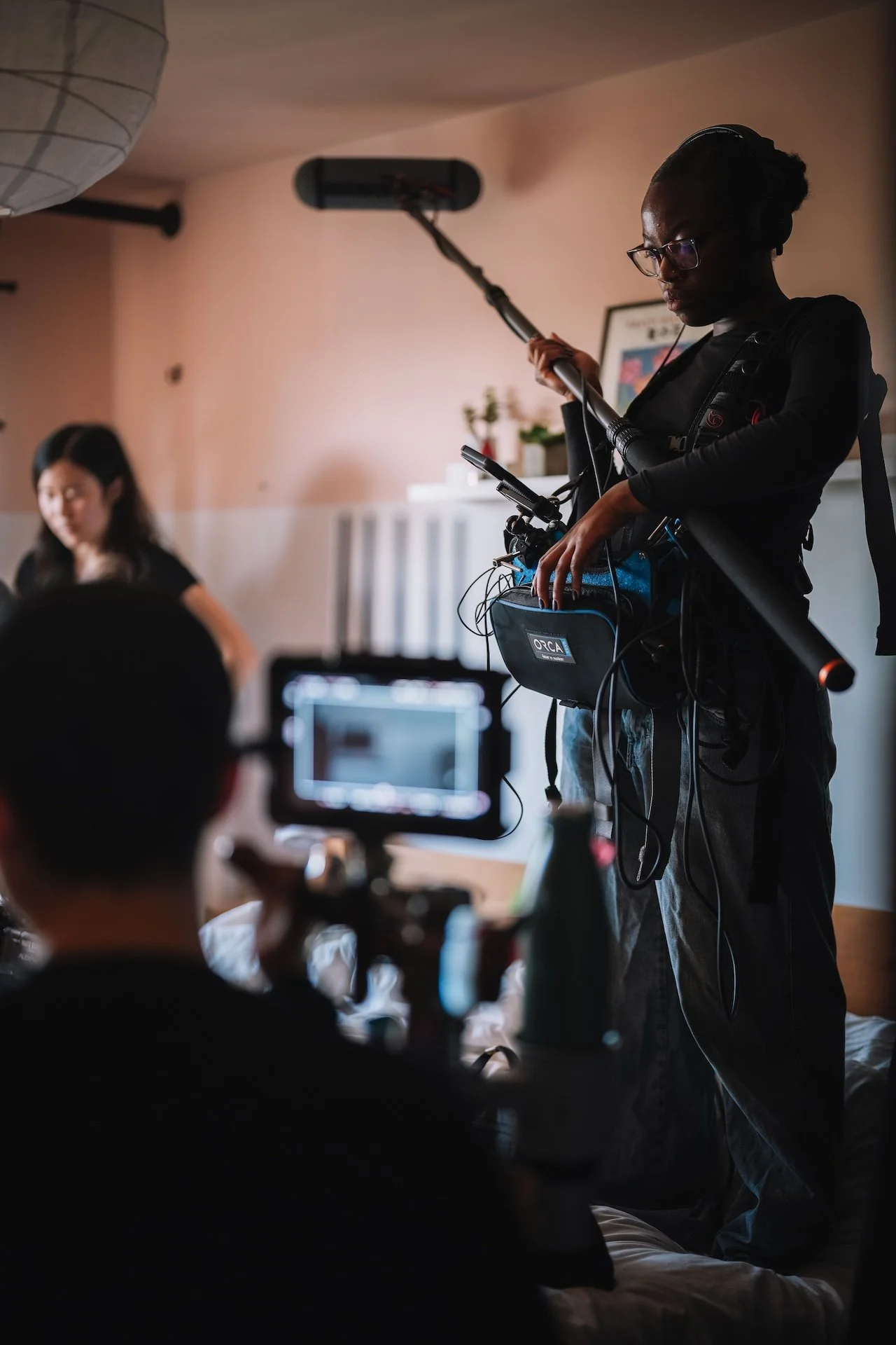 Filmmaker holding a boom microphone on a film set with a woman in the background, camera operator capturing scene.