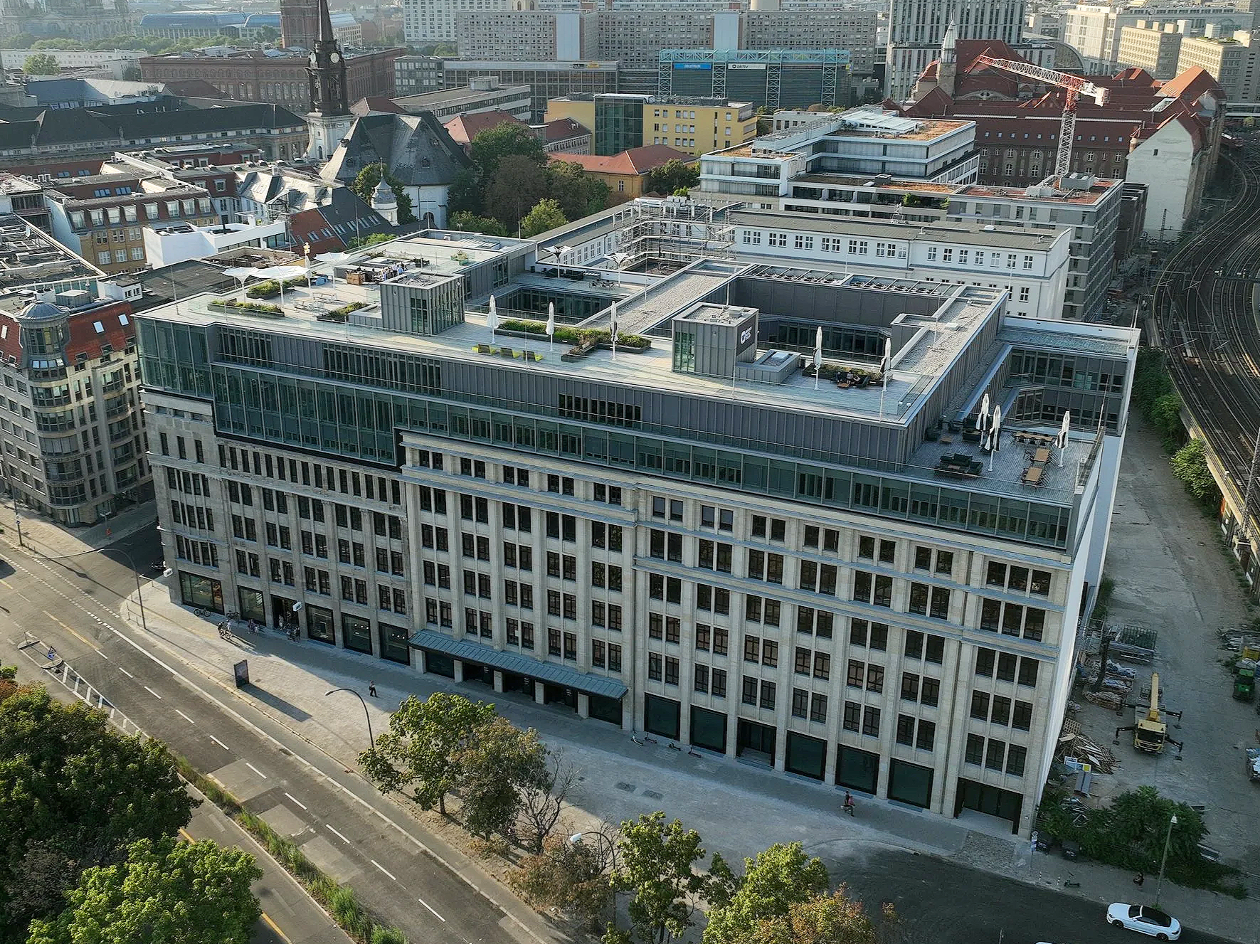Aerial view of a multi-story modern office building with a rooftop terrace, located in an urban area with roads, trees, and other buildings surrounding it.