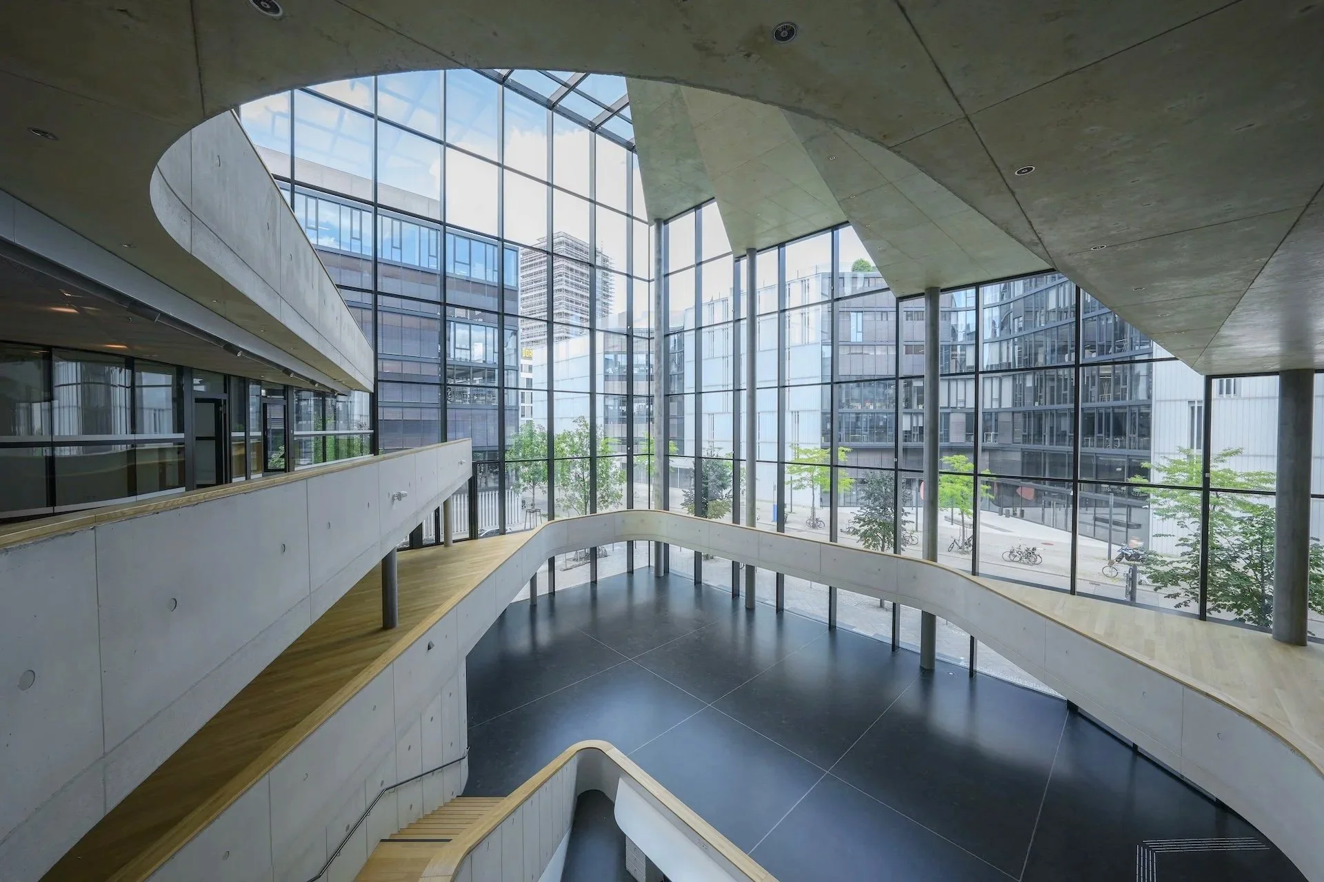 Interior view of a modern building with large glass windows, multiple levels with wooden walkways, and an urban cityscape outside.