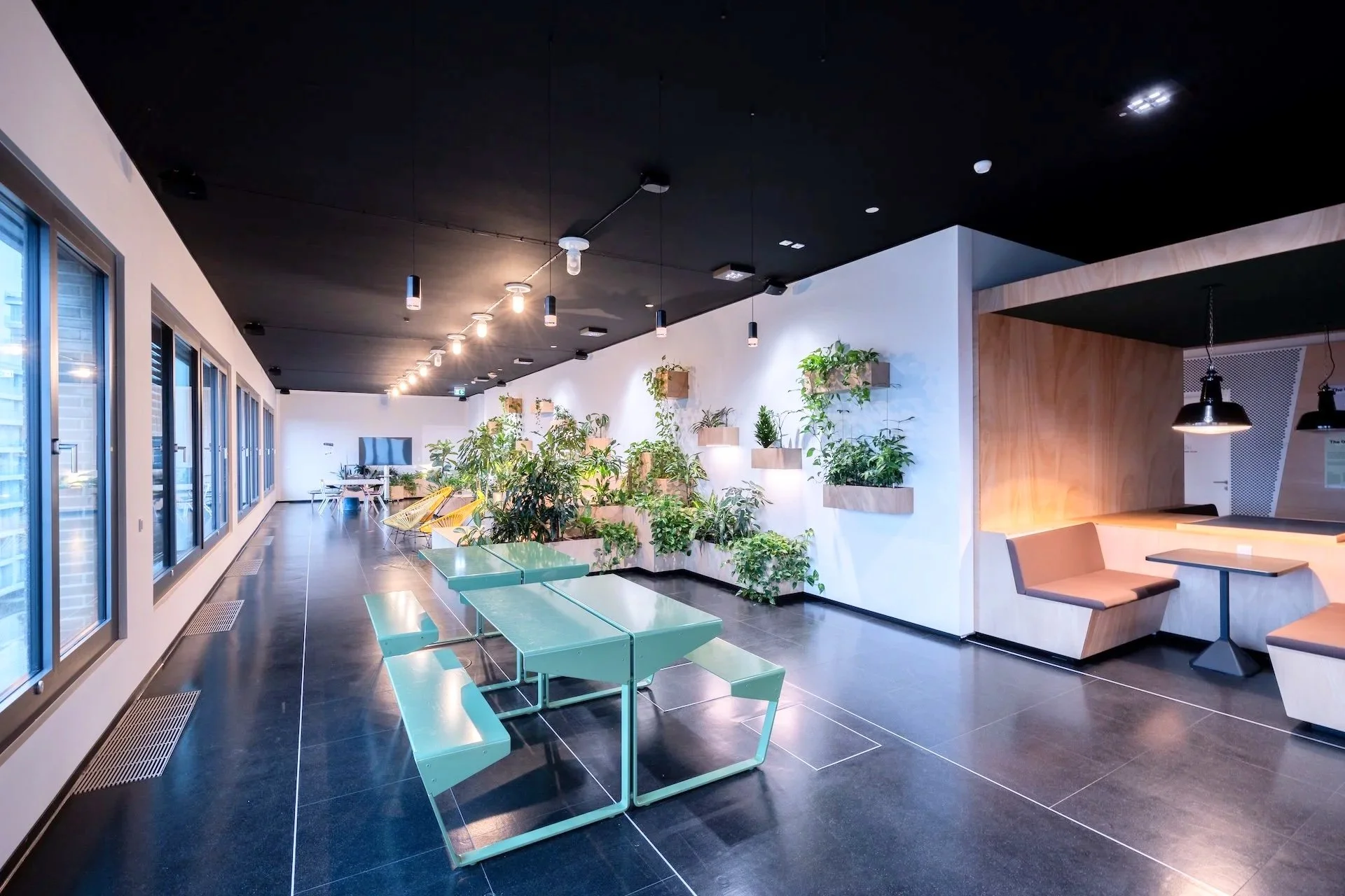 Modern indoor space with green plants arranged on white walls, tables, and seating areas, illuminated by overhead lights.