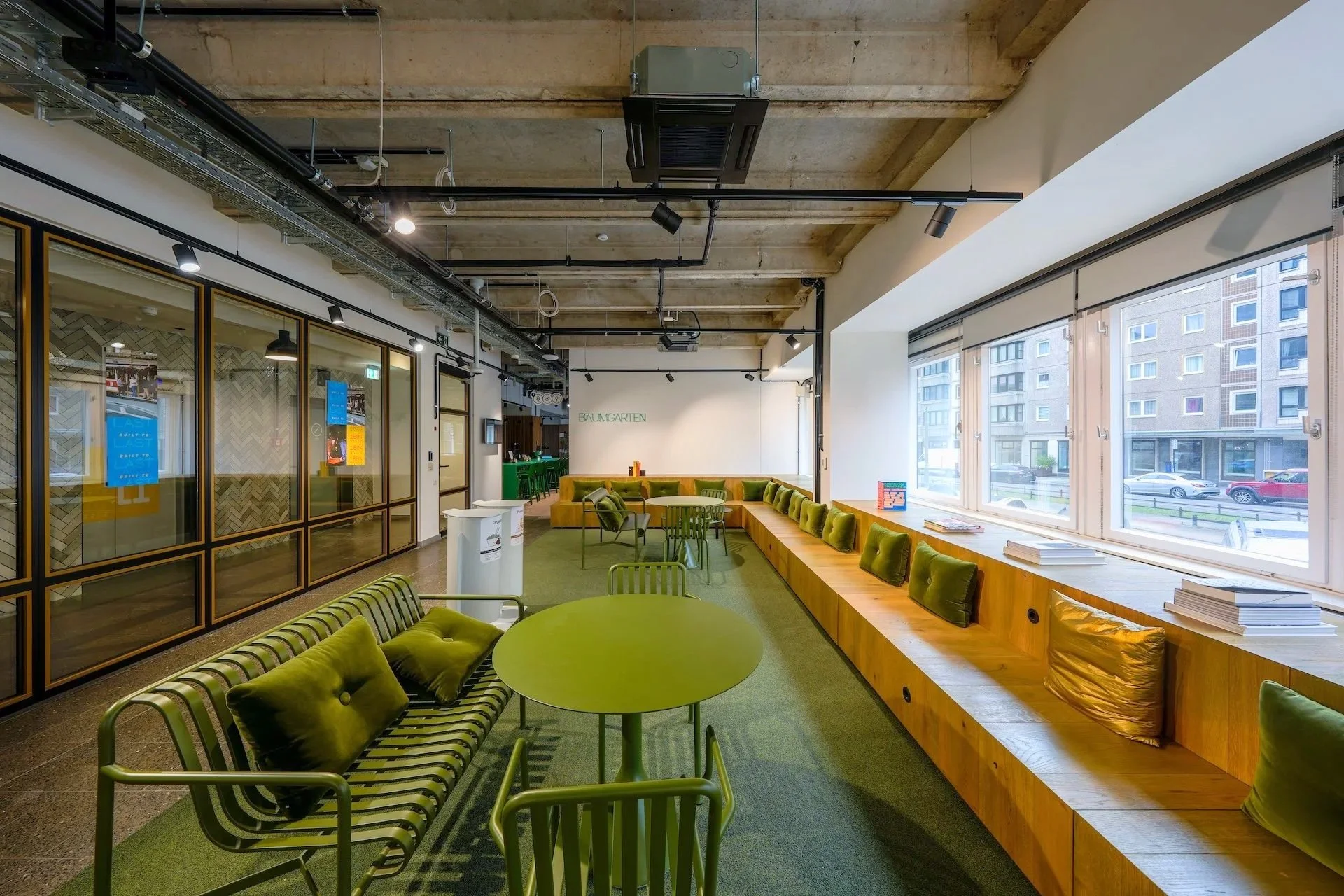 Modern office lounge with green chairs, a wooden bench with cushions by large windows, and a round table with chairs. Exposed ceiling with lighting and ductwork.