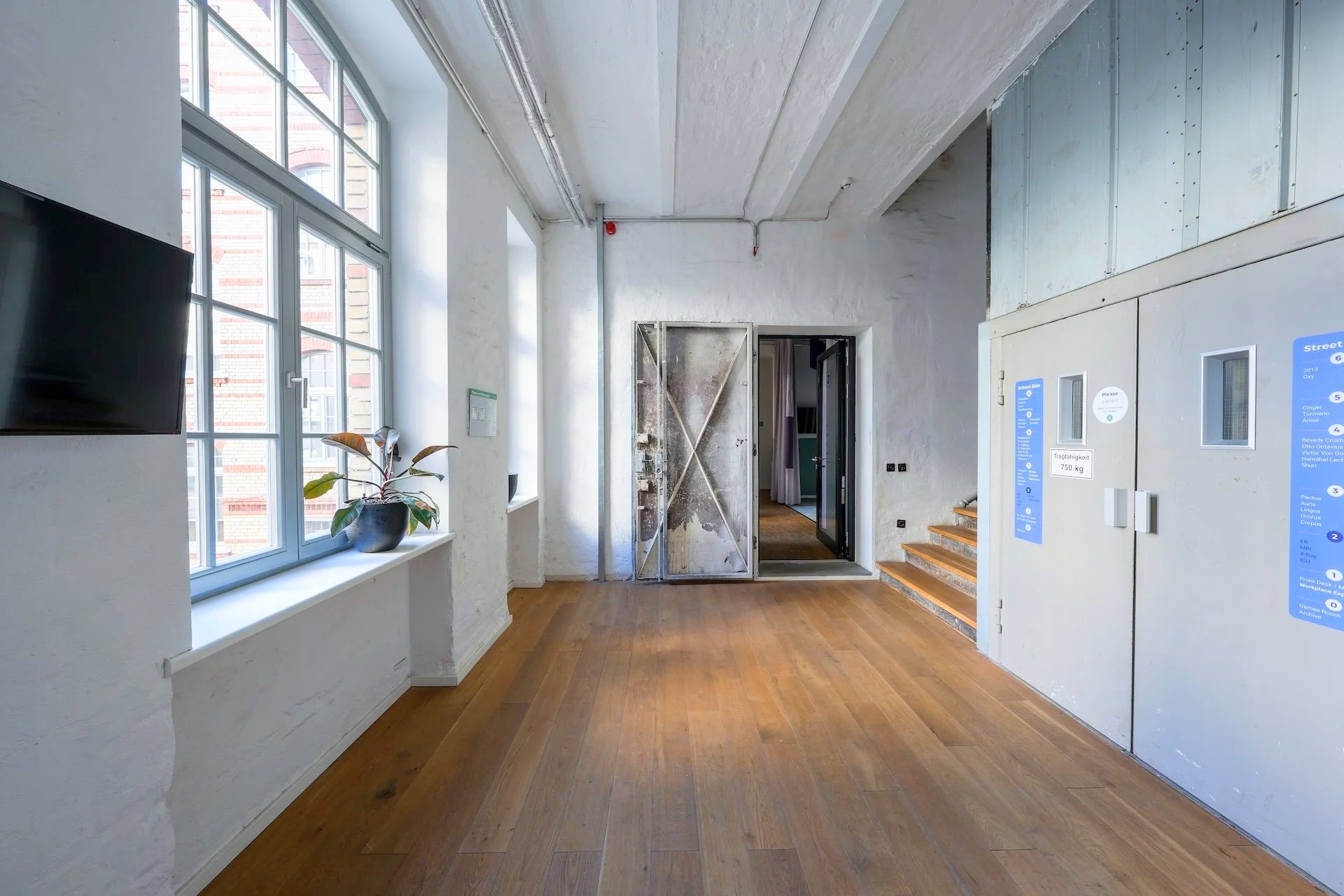 Interior of a building with wooden floor, white walls, large windows, a potted plant on the windowsill, and an open metal door at the back.